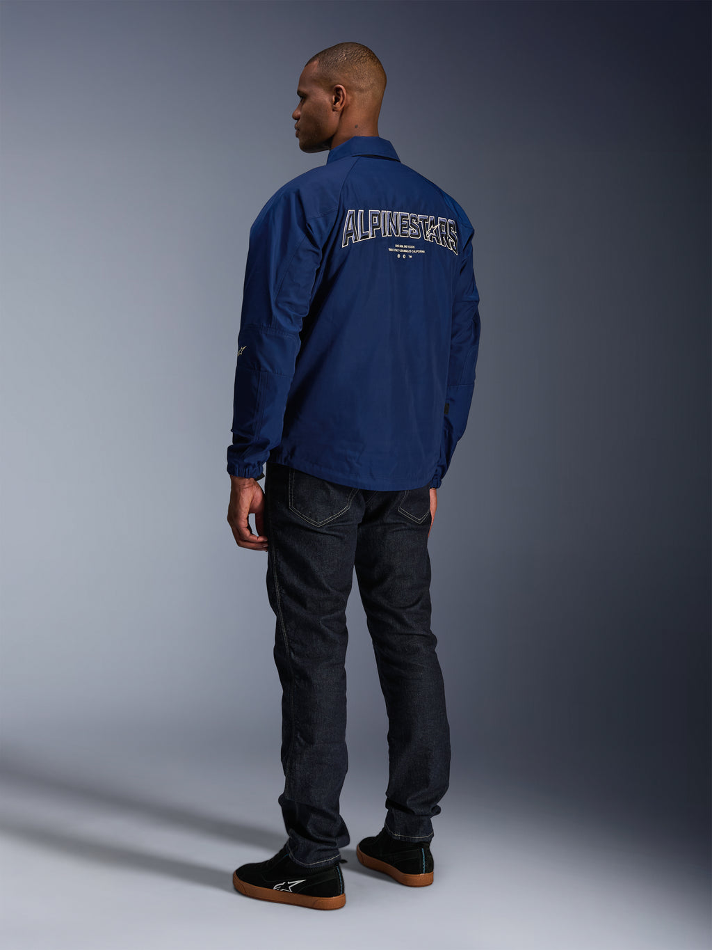 Model w kurtce Alpinestars Revved WR w kolorze granatowym, widok z tyłu, duży haft 'ALPINESTARS' z białym obramowaniem na plecach, ciemne spodnie jeansowe i czarne sneakersy, miejski styl motocyklowy