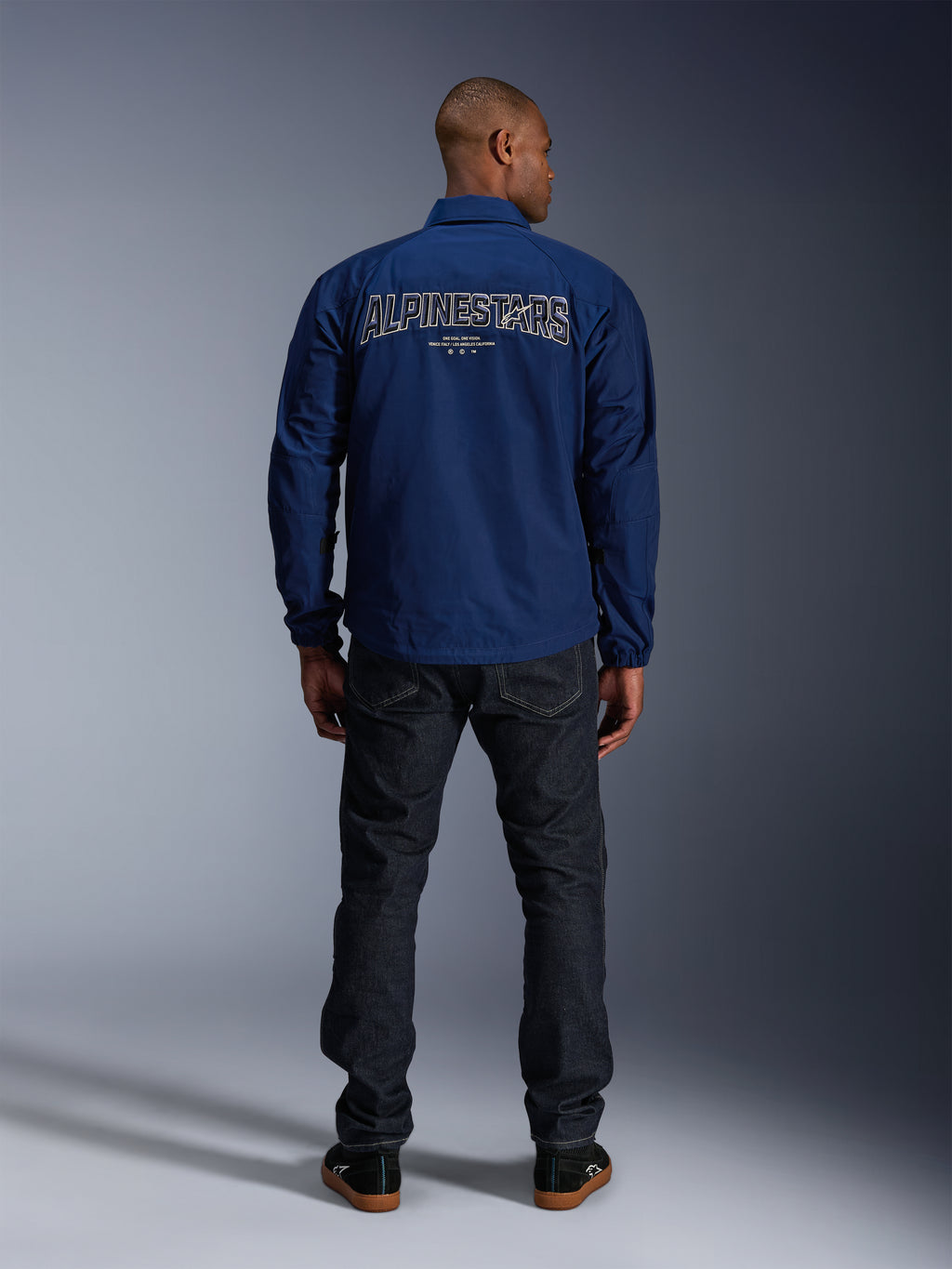 Kurtka Alpinestars Revved WR w kolorze granatowym, widok z tyłu, z dużym haftem „ALPINESTARS” z efektem 3D na plecach, stylizowana z ciemnymi dżinsami i czarnymi sneakersami, miejska odzież motocyklowa.