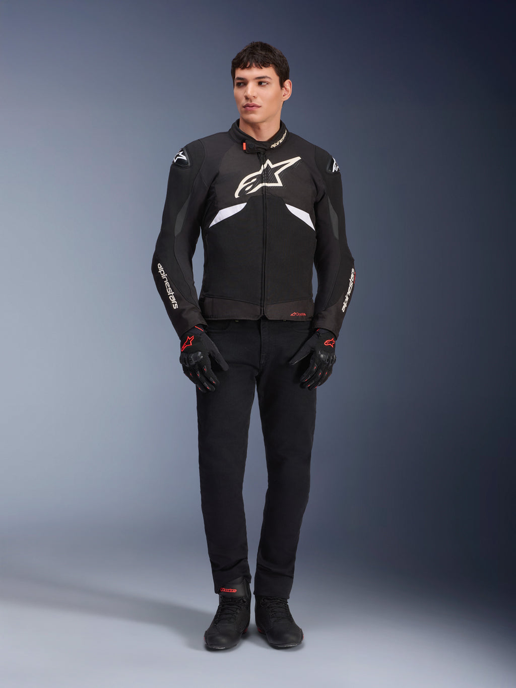 Male model wearing the Alpinestars T-GP R V3 Drystar® Jacket in Black and White, waterproof textile sport jacket with large white Alpinestars logo on the chest, worn with black riding pants and gloves, professional studio portrait highlighting the sleek sport silhouette.