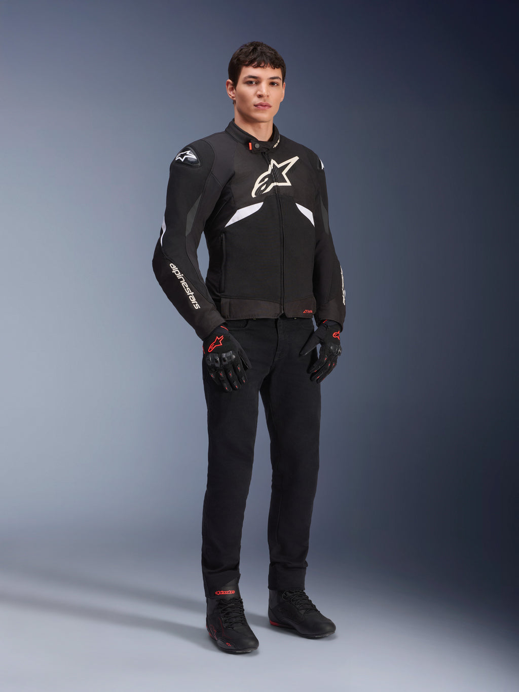Alpinestars T-GP R V3 Drystar® Jacket in Black and White, worn by a model with black motorcycle gloves and pants, featuring a white chest logo, white sleeve accents, and protective shoulder sliders, waterproof textile sport jacket for road riding