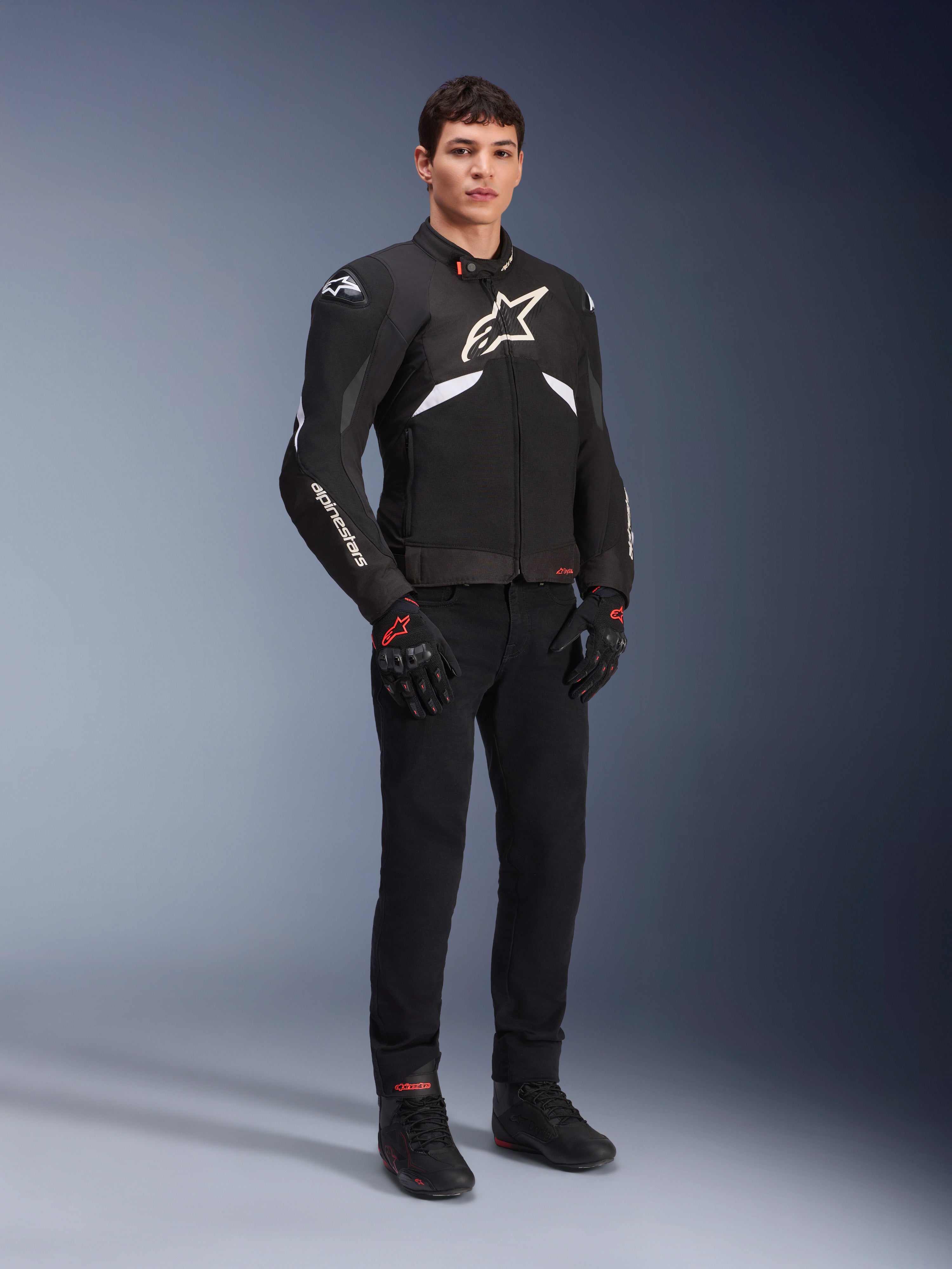 Alpinestars T-GP R V3 Drystar® Jacket in Black and White, worn by a model with black motorcycle gloves and pants, featuring a white chest logo, white sleeve accents, and protective shoulder sliders, waterproof textile sport jacket for road riding