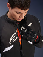Alpinestars T-GP R V3 Drystar® Jacket, Black and White motorcycle sport textile jacket, partially unzipped revealing orange 'Alpinestars Protects' inner strap, worn with black and red mesh gloves, race-derived protective apparel.
