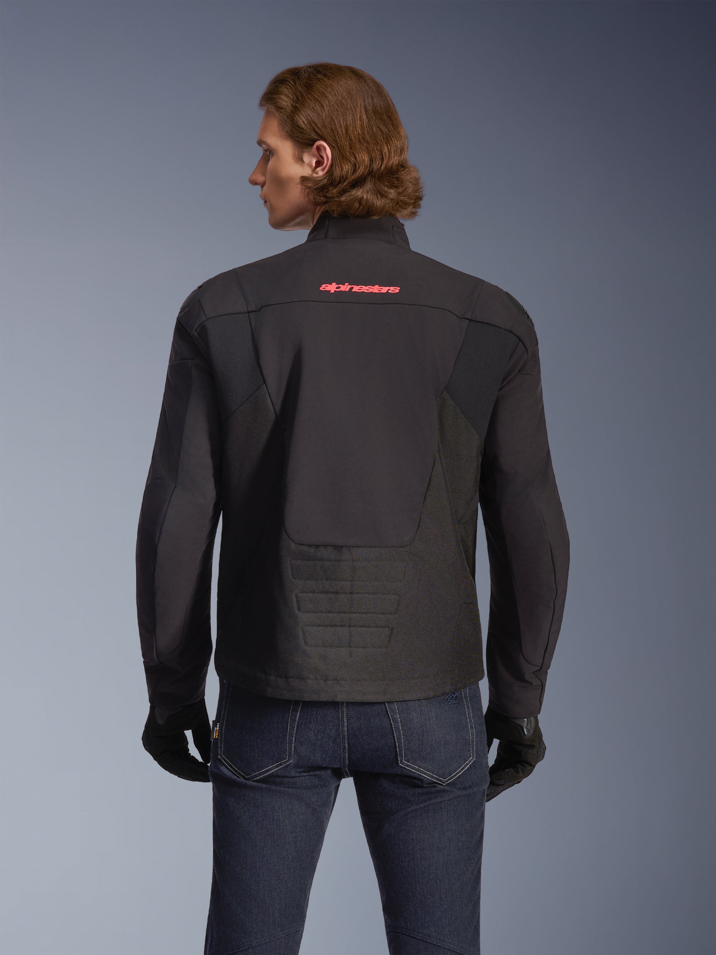 Alpinestars SMX Waterproof Jacket, czarna z jaskrawoczerwonym logo, widok z tyłu na modelu, noszona z ciemnoniebieskimi dżinsami i czarnymi rękawicami, tekstylna kurtka motocyklowa do jazdy szosowej i sportowej.