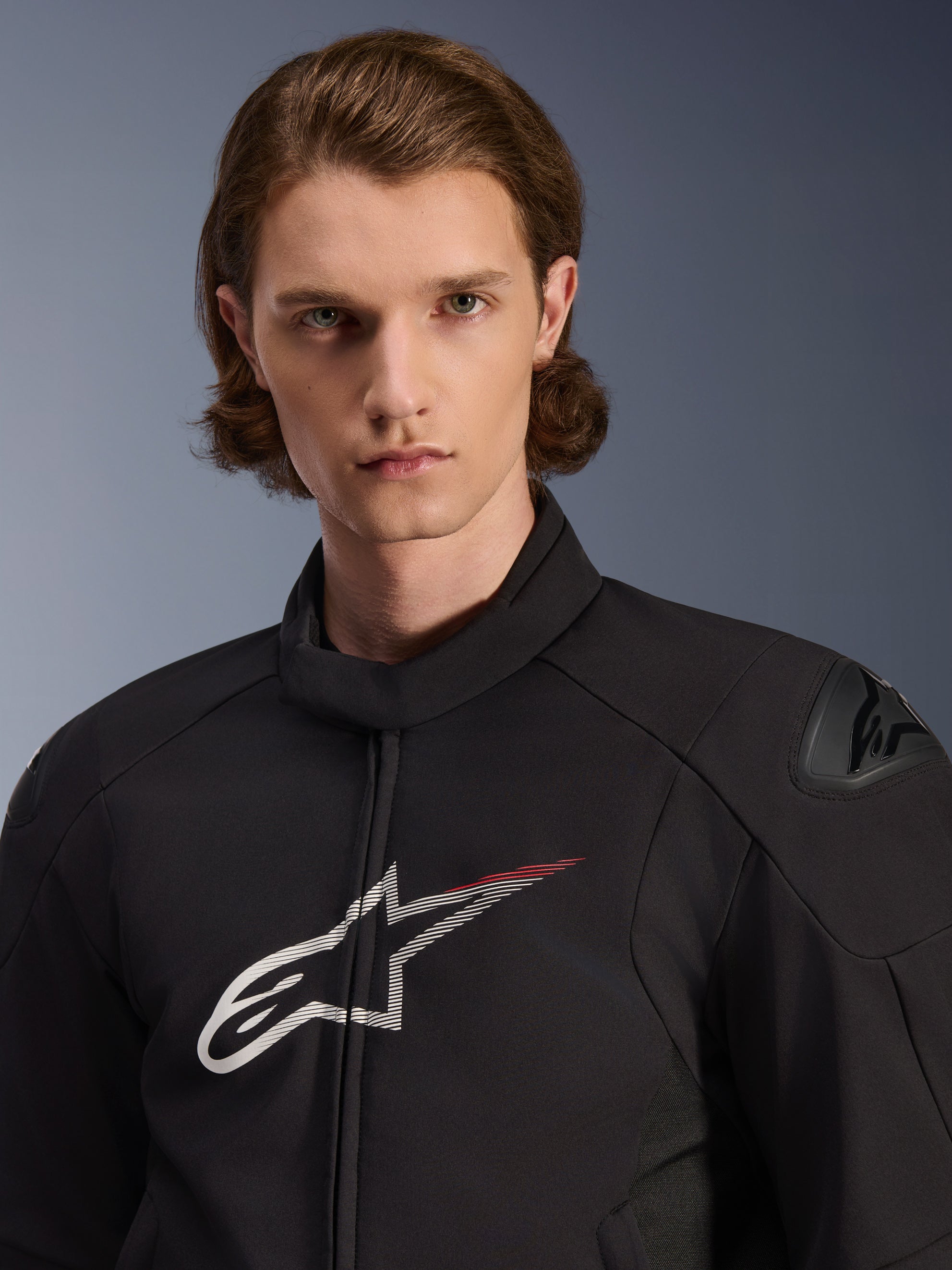 Model w kurtce Alpinestars SMX Waterproof Jacket, czarna z białymi i jaskrawoczerwonymi akcentami, duże białe logo gwiazdy z czerwonym paskiem na klatce piersiowej, ochraniacze ramion DFS Lite, nowoczesna konstrukcja tekstylna zapewniająca wydajność na drodze