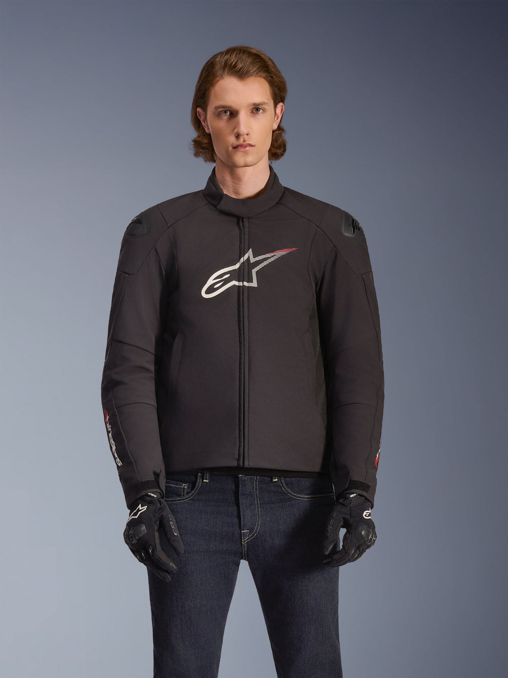 Alpinestars SMX Waterproof Jacket, czarna tekstylna kurtka motocyklowa z biało-czerwonym logo gwiazdy na piersi, noszona przez modela w czarnych rękawicach i niebieskich jeansach, wyposażona w zewnętrzne slidery na ramionach i sportowy krój.