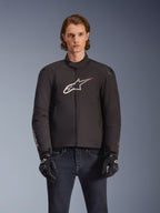 Alpinestars SMX Waterproof Jacket, czarna tekstylna kurtka motocyklowa z biało-czerwonym logo gwiazdy na piersi, noszona przez modela w czarnych rękawicach i niebieskich jeansach, wyposażona w zewnętrzne slidery na ramionach i sportowy krój.