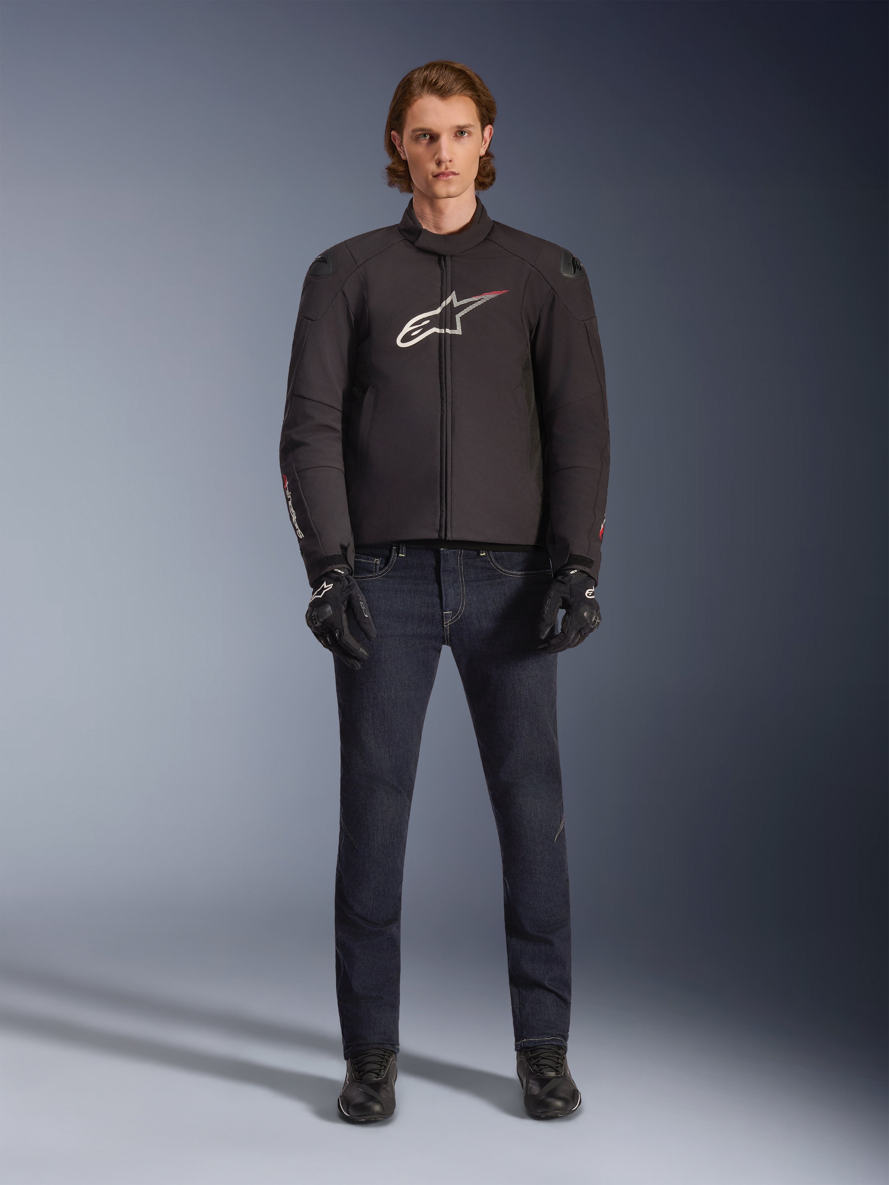 Model ubrany w kurtkę Alpinestars SMX Waterproof Jacket w kolorze czarnym z biało-czerwonym logo, w zestawieniu z ciemnymi jeansowymi spodniami motocyklowymi, czarnymi rękawicami i butami motocyklowymi, sportowy zestaw odzieży motocyklowej na każdą pogodę.