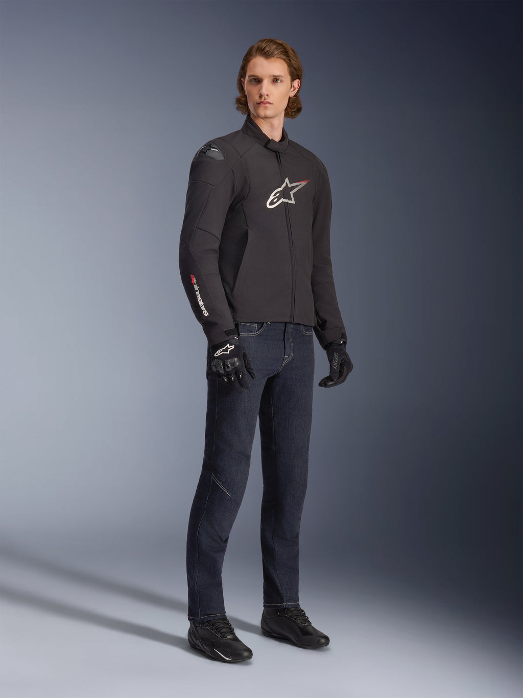 Męski model ubrany w wodoodporną kurtkę Alpinestars SMX, czarną z białymi i czerwonymi akcentami logo, zestawioną z ciemnoniebieskimi jeansami i czarnymi rękawicami motocyklowymi, smukła tekstylna kurtka motocyklowa o sportowym kroju.