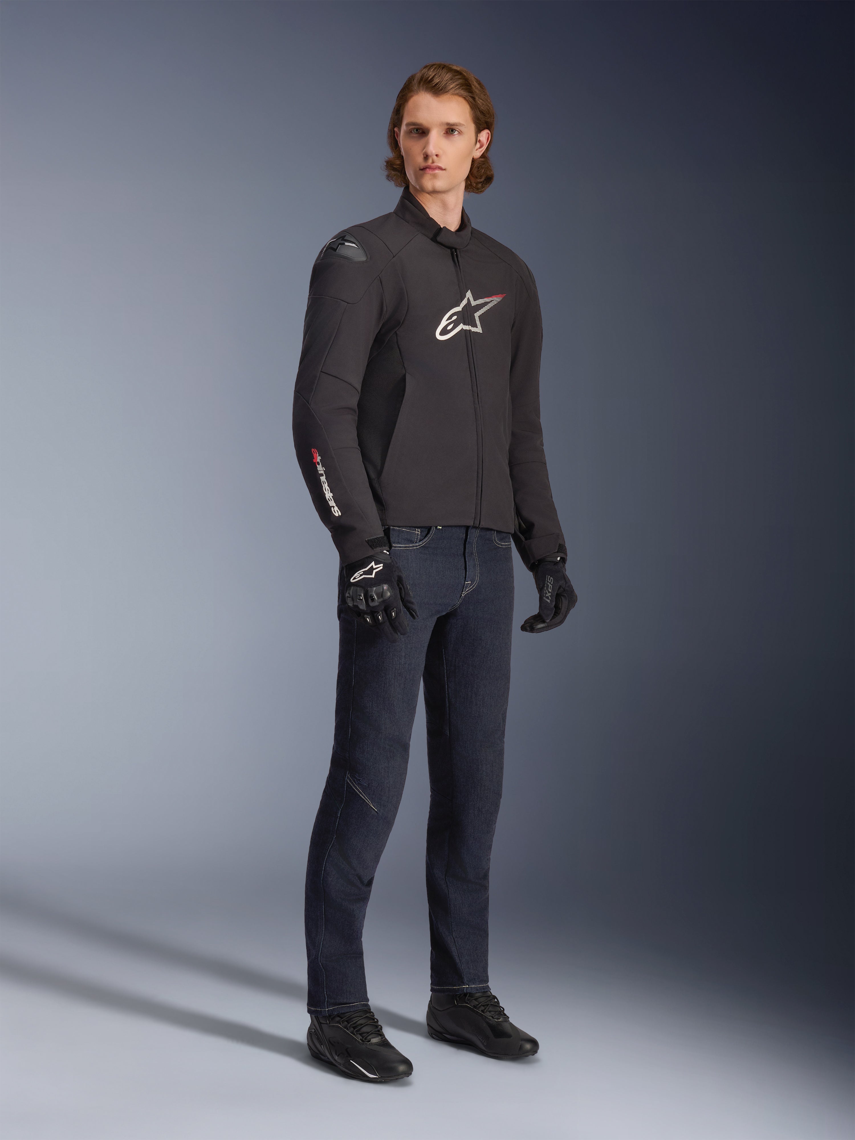 Męski model ubrany w wodoodporną kurtkę Alpinestars SMX, czarną z białymi i czerwonymi akcentami logo, zestawioną z ciemnoniebieskimi jeansami i czarnymi rękawicami motocyklowymi, smukła tekstylna kurtka motocyklowa o sportowym kroju.
