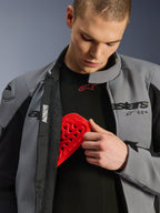 Męski model w wodoodpornej kurtce Alpinestars SMX w kolorze Dark Gray Black, wkładający czerwony ochraniacz klatki piersiowej Nucleon do wewnętrznej kieszeni; prezentacja kompatybilności kurtki z ochraniaczami i jej funkcji ochronnych.