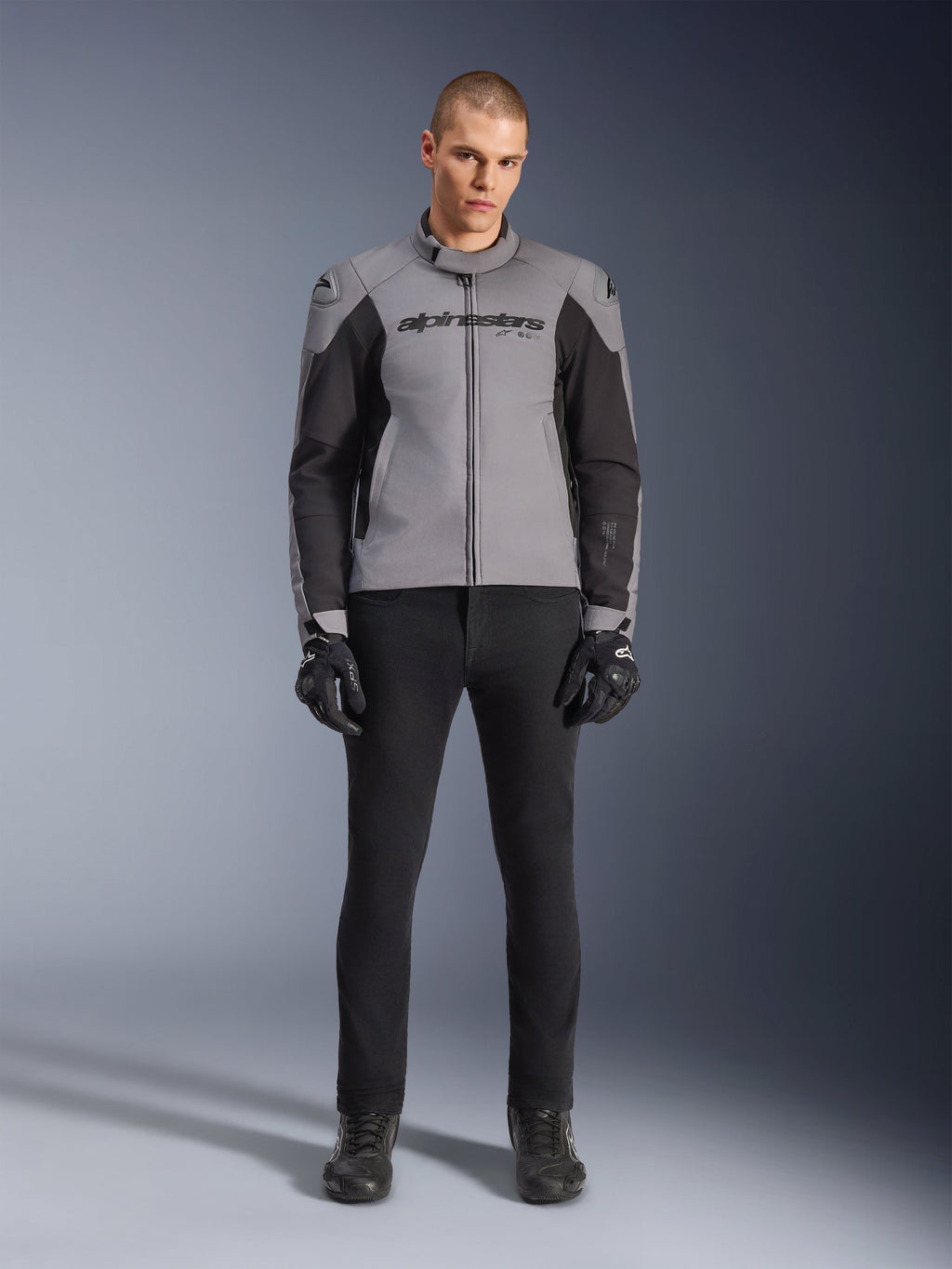 Kurtka Alpinestars SMX Waterproof, ciemnoszara i czarna, widok z przodu na modelu, tekstylna sportowa kurtka motocyklowa z czarnymi panelami bocznymi i logo Alpinestars na klatce piersiowej, w zestawieniu z czarnymi spodniami i rękawicami motocyklowymi.