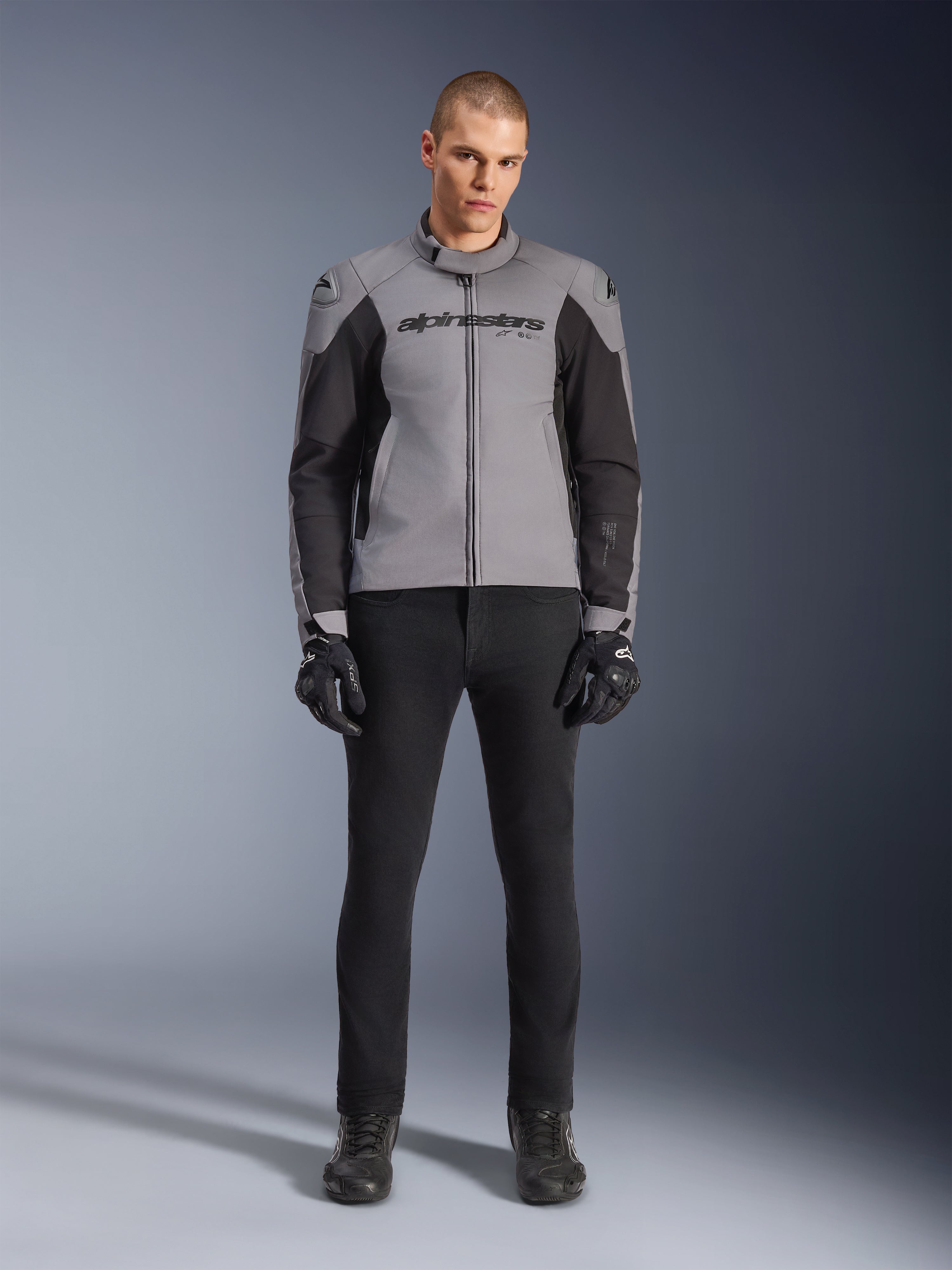 Kurtka Alpinestars SMX Waterproof, ciemnoszara i czarna, widok z przodu na modelu, tekstylna sportowa kurtka motocyklowa z czarnymi panelami bocznymi i logo Alpinestars na klatce piersiowej, w zestawieniu z czarnymi spodniami i rękawicami motocyklowymi.