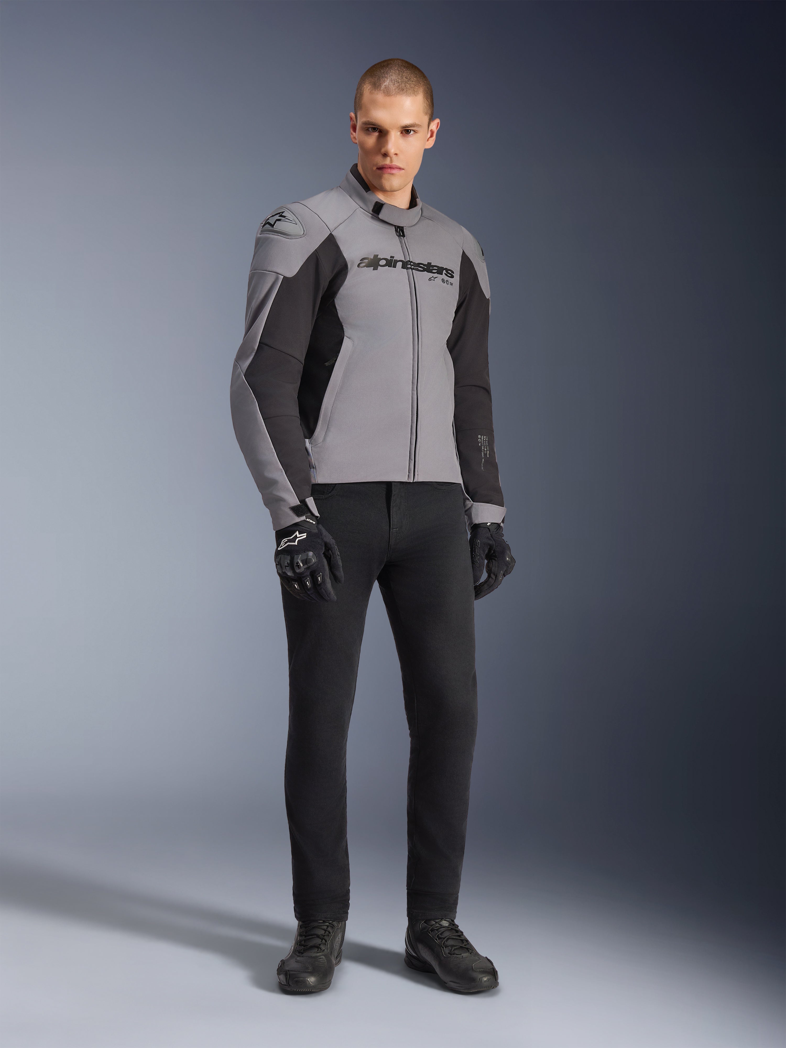 Alpinestars SMX Waterproof Jacket, kurtka motocyklowa, ciemnoszara i czarna, noszona przez modela z czarnymi rękawicami i spodniami, posiada wyraźne logo na klatce piersiowej i techniczny sportowy krój do jazdy szosowej w każdych warunkach pogodowych.