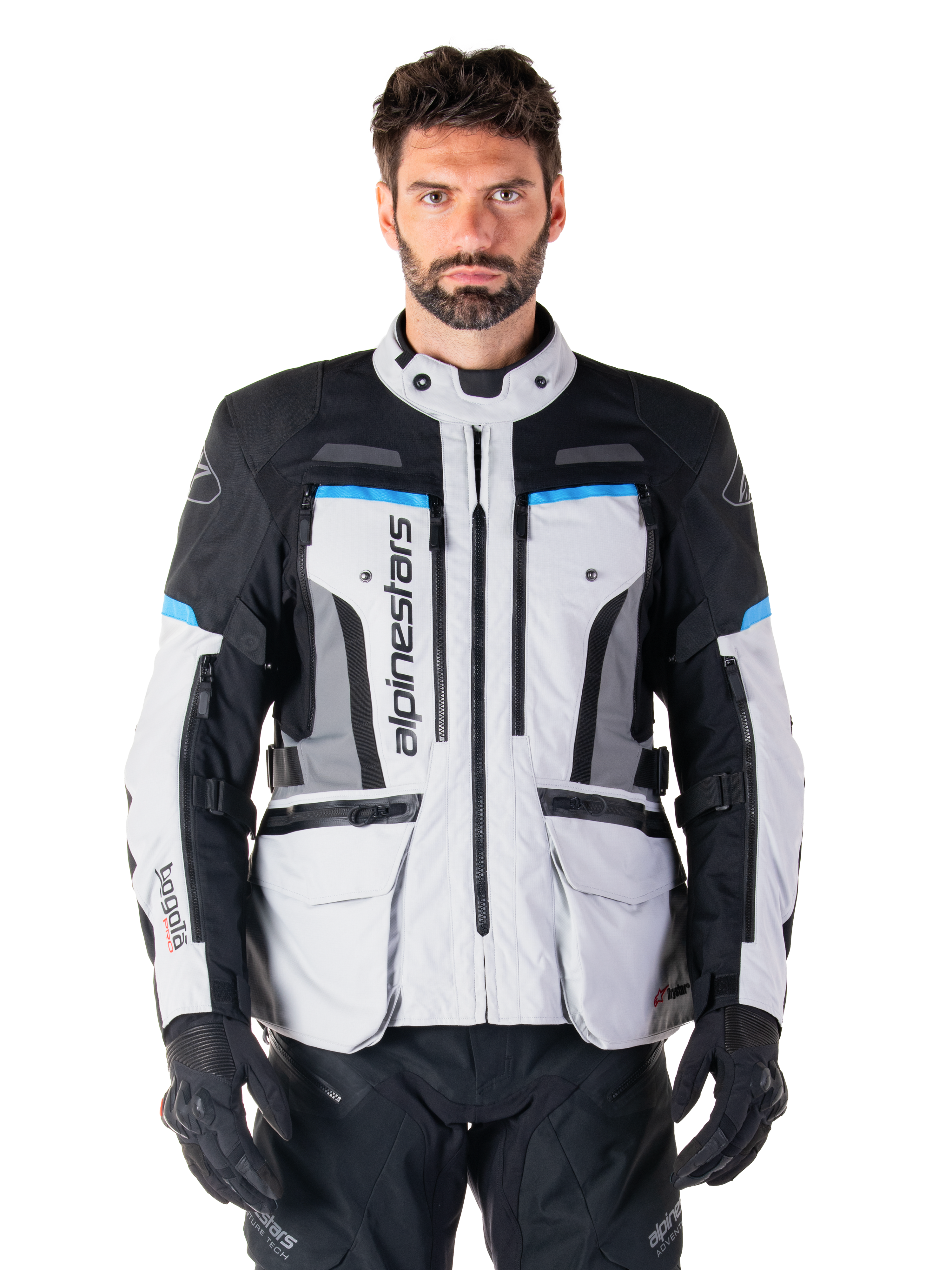Alpinestars Bogota Pro Drystar Jacket, motocyklowa kurtka turystyczna adventure, kolor Ice Gray z czarnymi i błękitnymi akcentami, widok z przodu ukazujący panele wentylacyjne i kieszenie cargo, model w czarnych spodniach i rękawicach