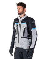 Kurtka Alpinestars Bogota' Pro Drystar®, kolor jasnoszary, czarny i cyjanowy, motocyklowa kurtka adventure noszona przez mężczyznę w czarnych rękawicach, techniczny design z licznymi kieszeniami i panelami wentylacyjnymi, wysokowydajna odzież motocyklowa