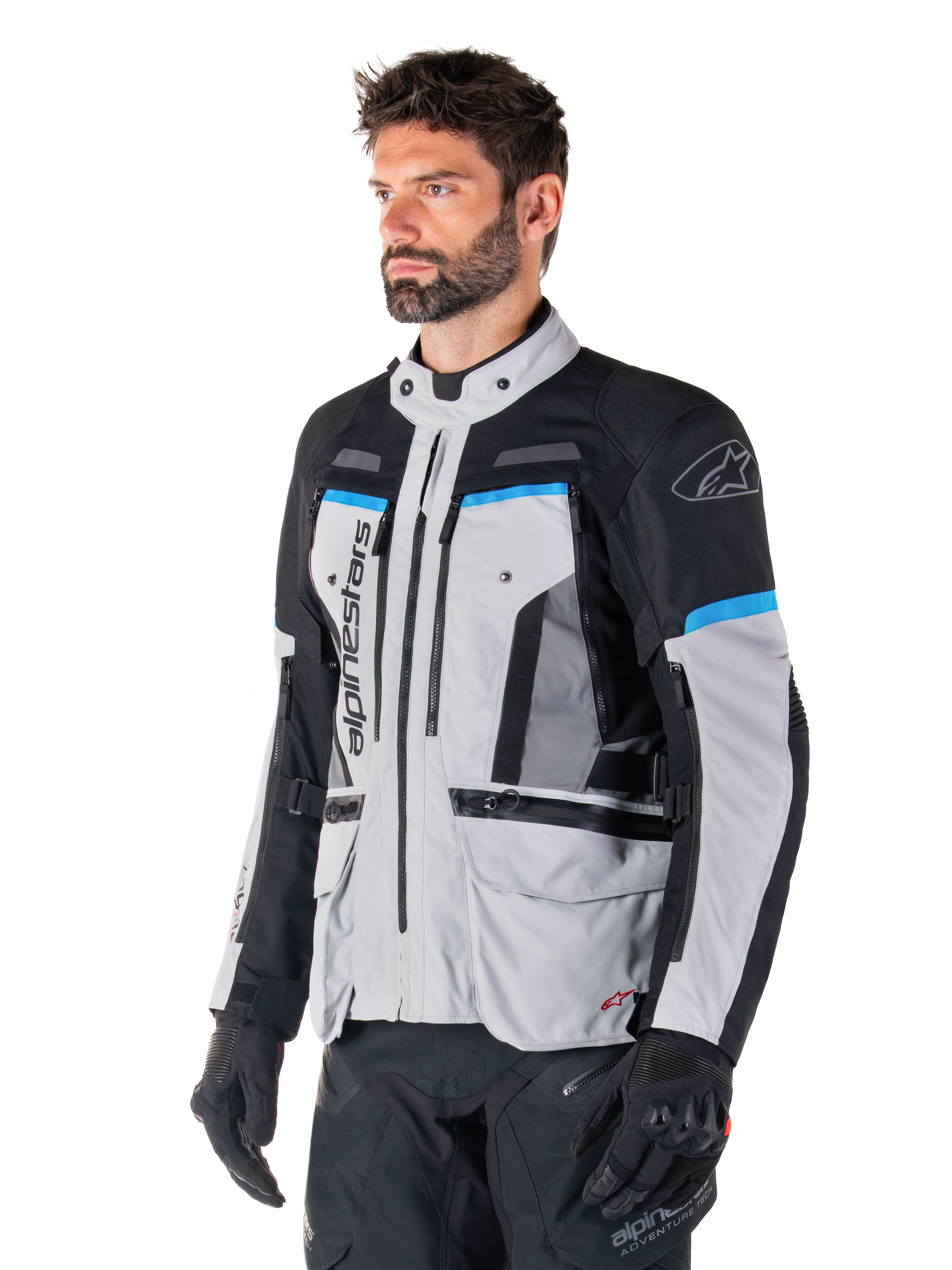 Kurtka Alpinestars Bogota' Pro Drystar®, kolor jasnoszary, czarny i cyjanowy, motocyklowa kurtka adventure noszona przez mężczyznę w czarnych rękawicach, techniczny design z licznymi kieszeniami i panelami wentylacyjnymi, wysokowydajna odzież motocyklowa