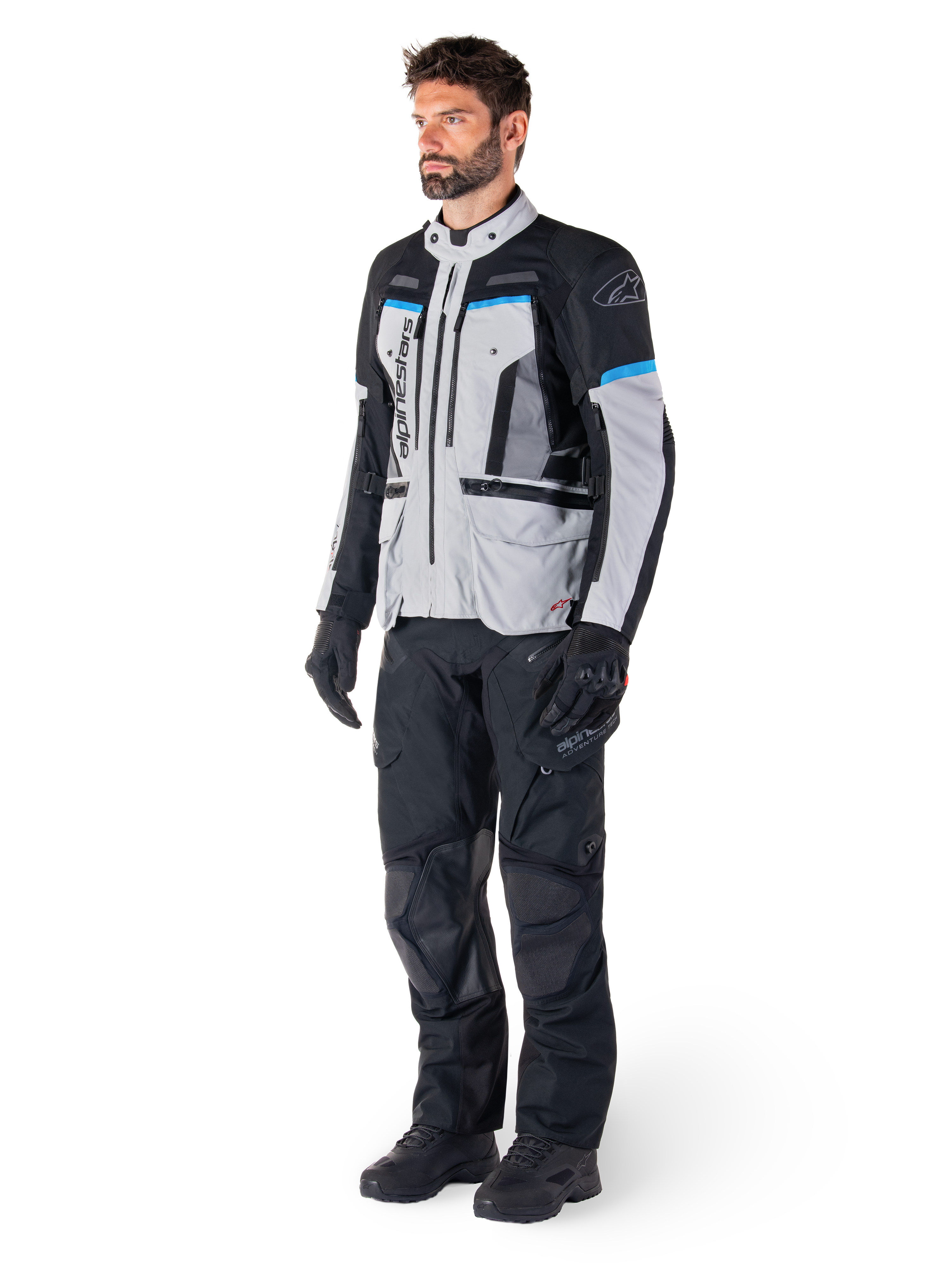 Motocyklista w kurtce Alpinestars Bogotá Pro Drystar® w kolorach Ice Gray, czarnym i cyjanowym, w zestawieniu z czarnymi technicznymi spodniami i rękawicami, odzież adventure touring zapewniająca wydajność w każdych warunkach pogodowych.