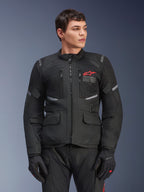 Alpinestars Andes Air Drystar® Jacket, czarna motocyklowa kurtka turystyczna, czerwone logo z gwiazdą na klatce piersiowej, panele wentylacyjne z siatki i odblaskowe akcenty, noszona przez męskiego modela w czarnych rękawicach, dwusezonowa odzież adventure