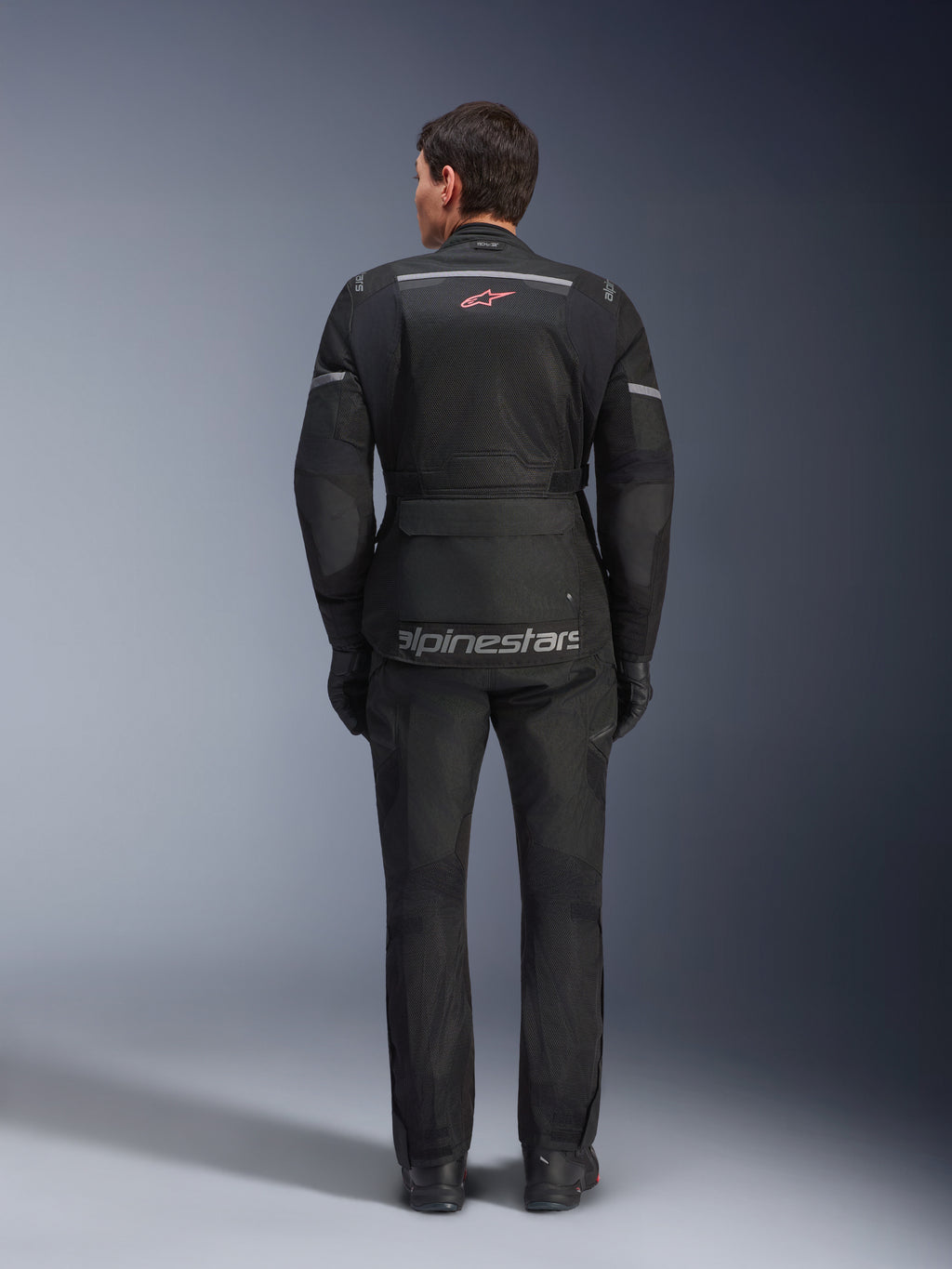 Widok z tyłu motocyklisty ubranego w kurtkę Alpinestars Andes Air Drystar® w kolorze czarnym, z rozległymi panelami wentylacyjnymi z siatki na plecach, czerwonym logo Alpinestars i odblaskowymi akcentami, w zestawie z pasującymi spodniami motocyklowymi i rękawicami do turystyki adventure.