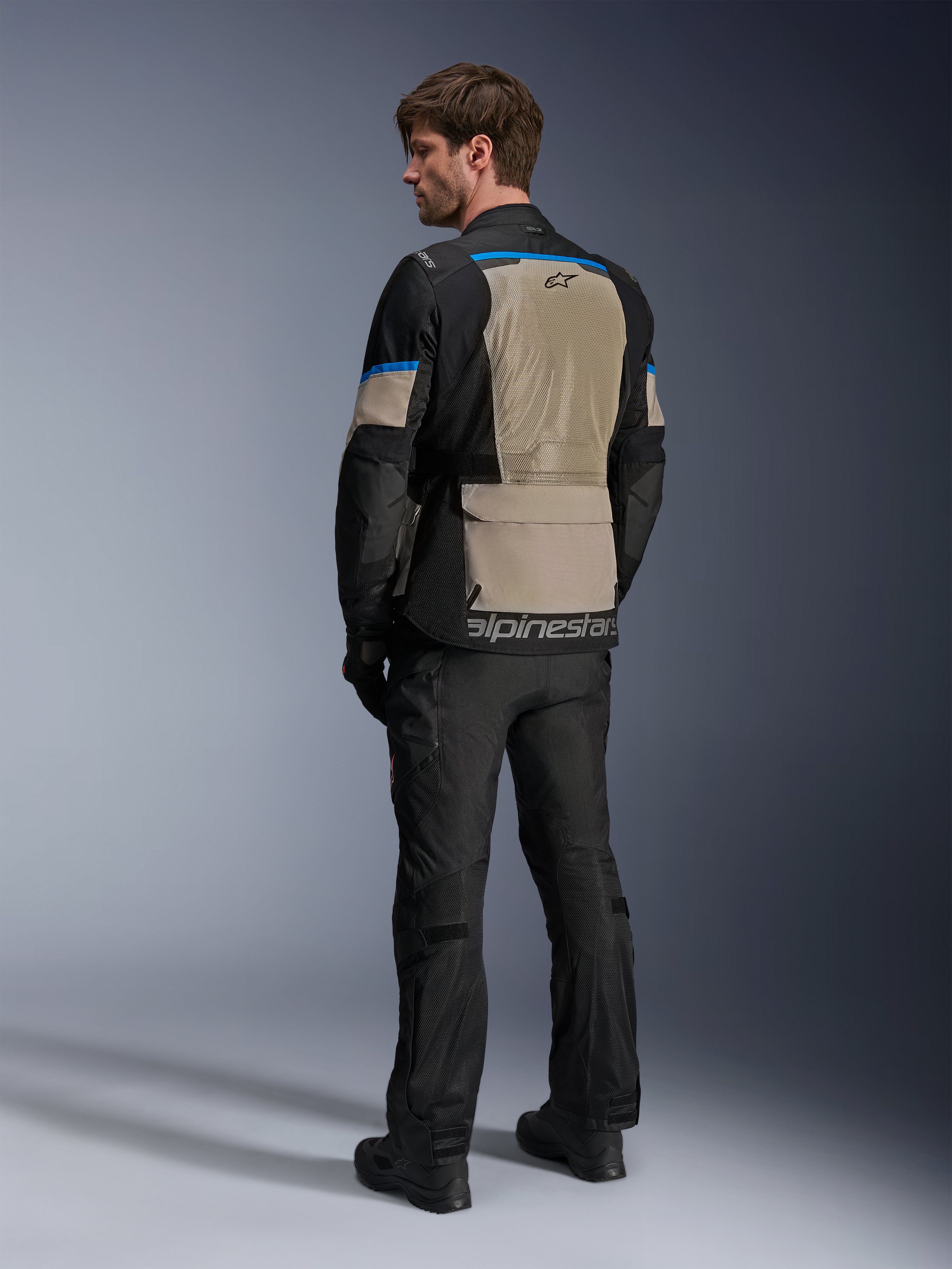 Kurtka Alpinestars Andes Air Drystar®, czarna, szara Laurel Gray i jasnoniebieska, widok z tyłu ukazujący duży siatkowy panel wentylacyjny na plecach, logo na dolnej krawędzi, w zestawie z czarnymi spodniami motocyklowymi i butami.
