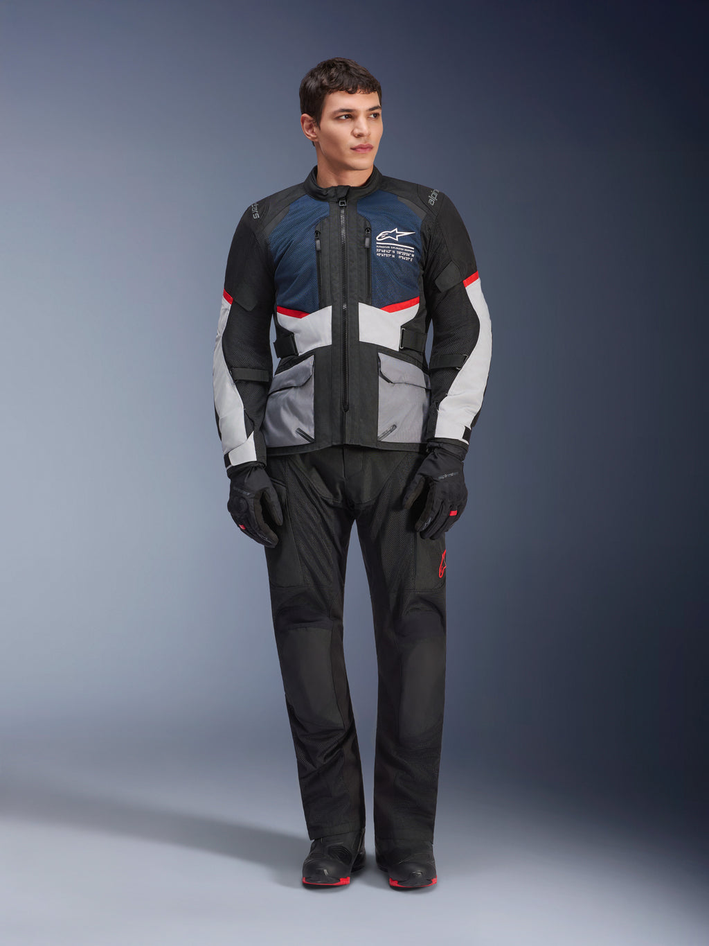 Kurtka Alpinestars Andes Air Drystar®, motocyklowa kurtka turystyczna, ciemnoniebieska, czarna i jasnoszara, noszona z pasującymi czarnymi spodniami adventure i rękawicami, panele z siatki dla przepływu powietrza, techniczna konstrukcja z wieloma kieszeniami.