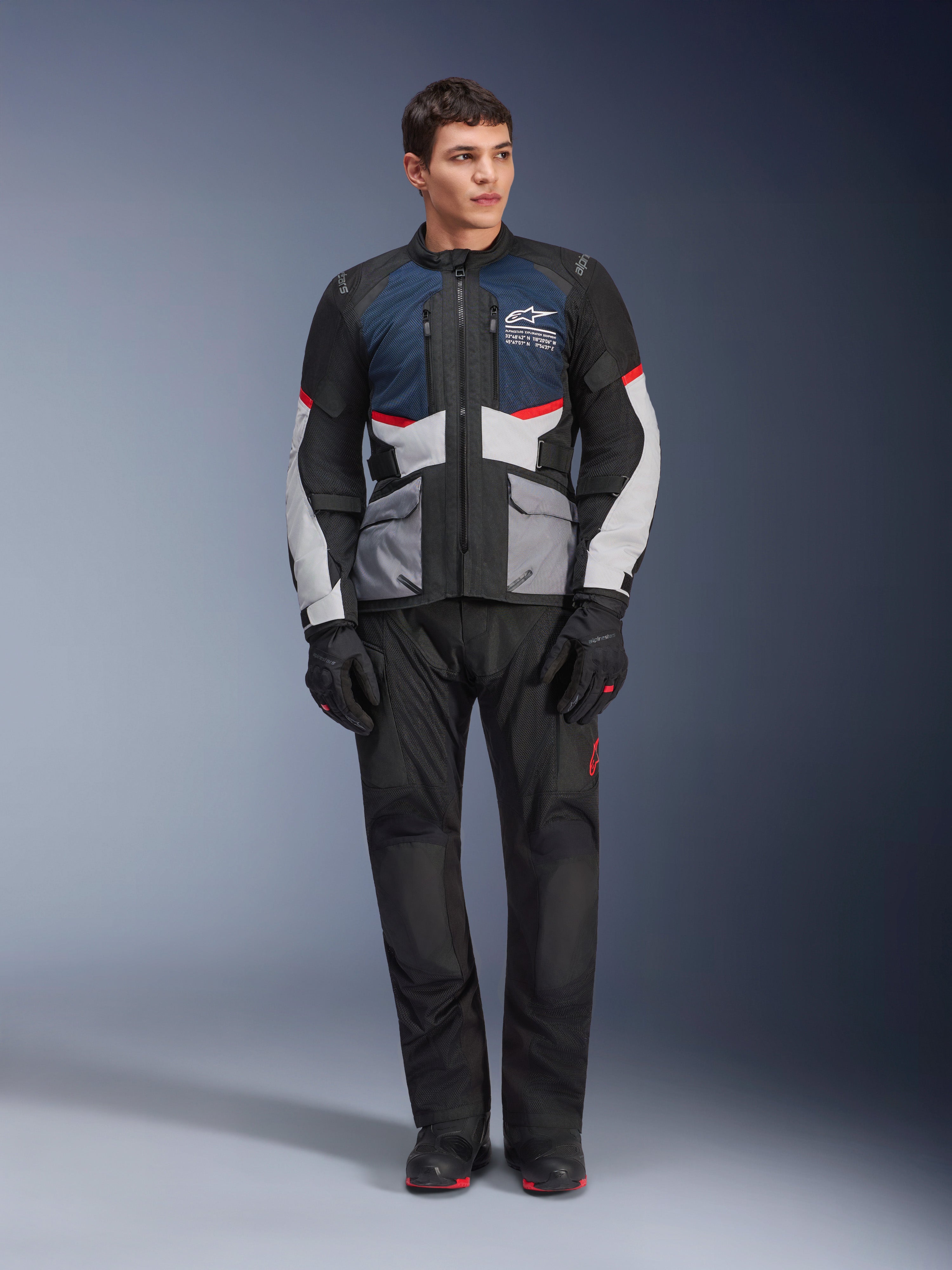 Kurtka Alpinestars Andes Air Drystar®, motocyklowa kurtka turystyczna, ciemnoniebieska, czarna i jasnoszara, noszona z pasującymi czarnymi spodniami adventure i rękawicami, panele z siatki dla przepływu powietrza, techniczna konstrukcja z wieloma kieszeniami.