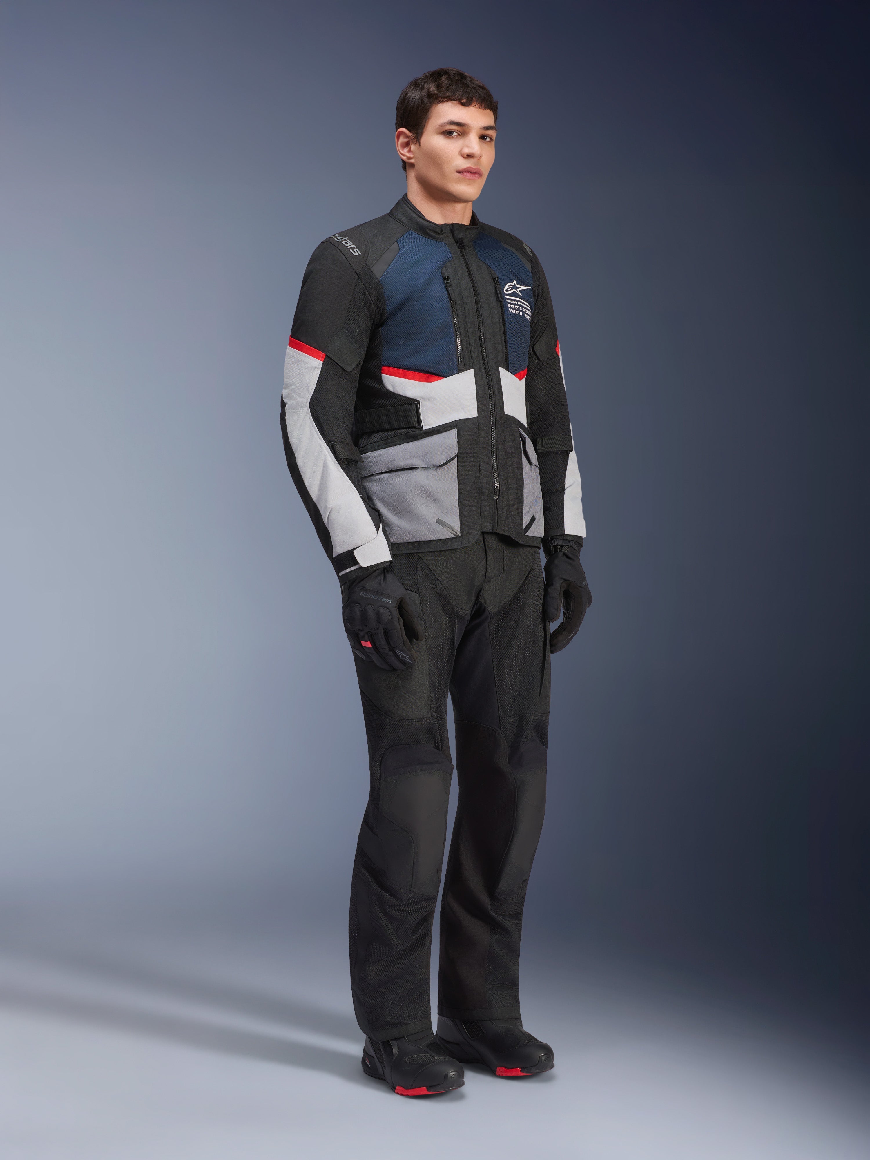 Męski model w kurtce Alpinestars Andes Air Drystar® w kolorze Deep Blue Black Ice Gray, w zestawieniu z czarnymi spodniami motocyklowymi, rękawicami i butami, z dużymi panelami siatkowymi dla wentylacji, jasnoszarymi kieszeniami i czerwonymi akcentami, przeznaczonej do turystyki adventure.