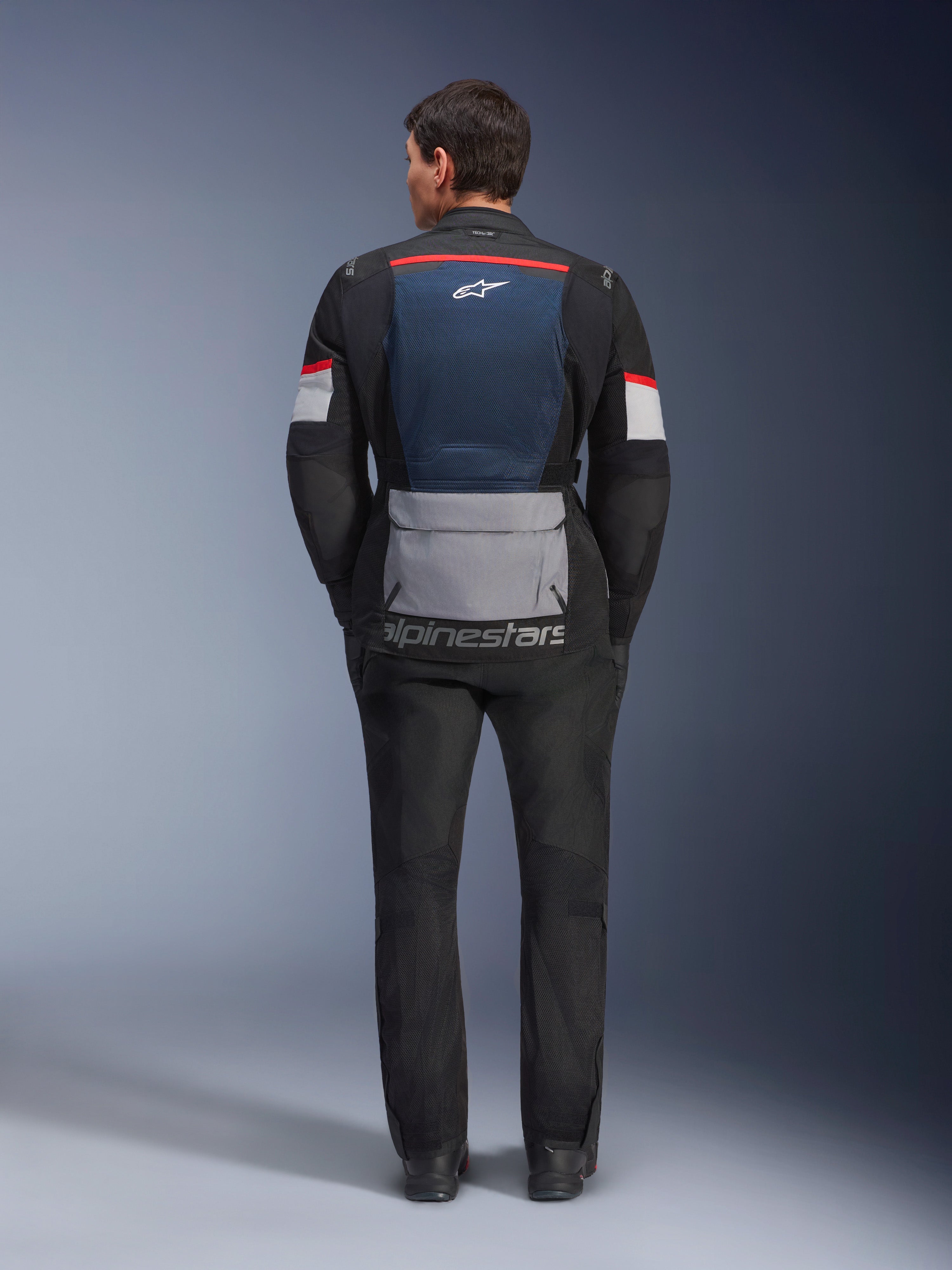 Kurtka Alpinestars Andes Air Drystar, Deep Blue Black Ice Gray, widok z tyłu na modelu, niebieski siatkowy panel wentylacyjny na plecach, duża szara kieszeń u dołu, czerwone i białe akcenty na rękawach, napis „alpinestars”, zestawiona z czarnymi spodniami motocyklowymi dla pełnego zestawu turystycznego