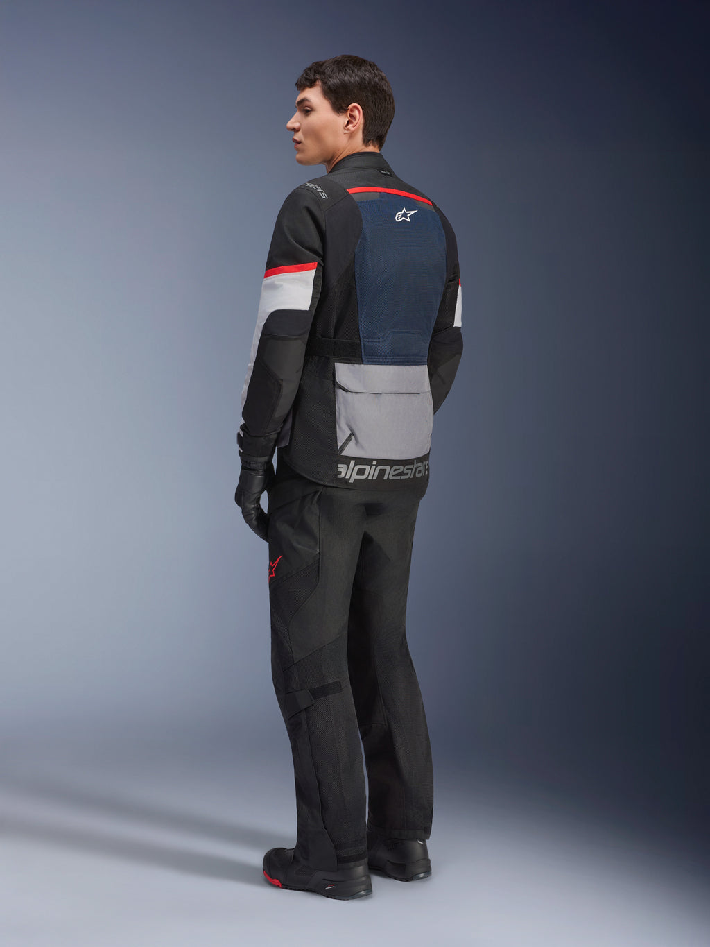 Męski model w ujęciu z tyłu i z boku, ubrany w kurtkę Alpinestars Andes Air Drystar® w kolorze Deep Blue Black Ice Gray, czarne spodnie motocyklowe i rękawice, niebieski panel siatkowy na plecach, szara kieszeń cargo na dole pleców i czerwone akcenty na rękawach, profesjonalna odzież adventure touring.
