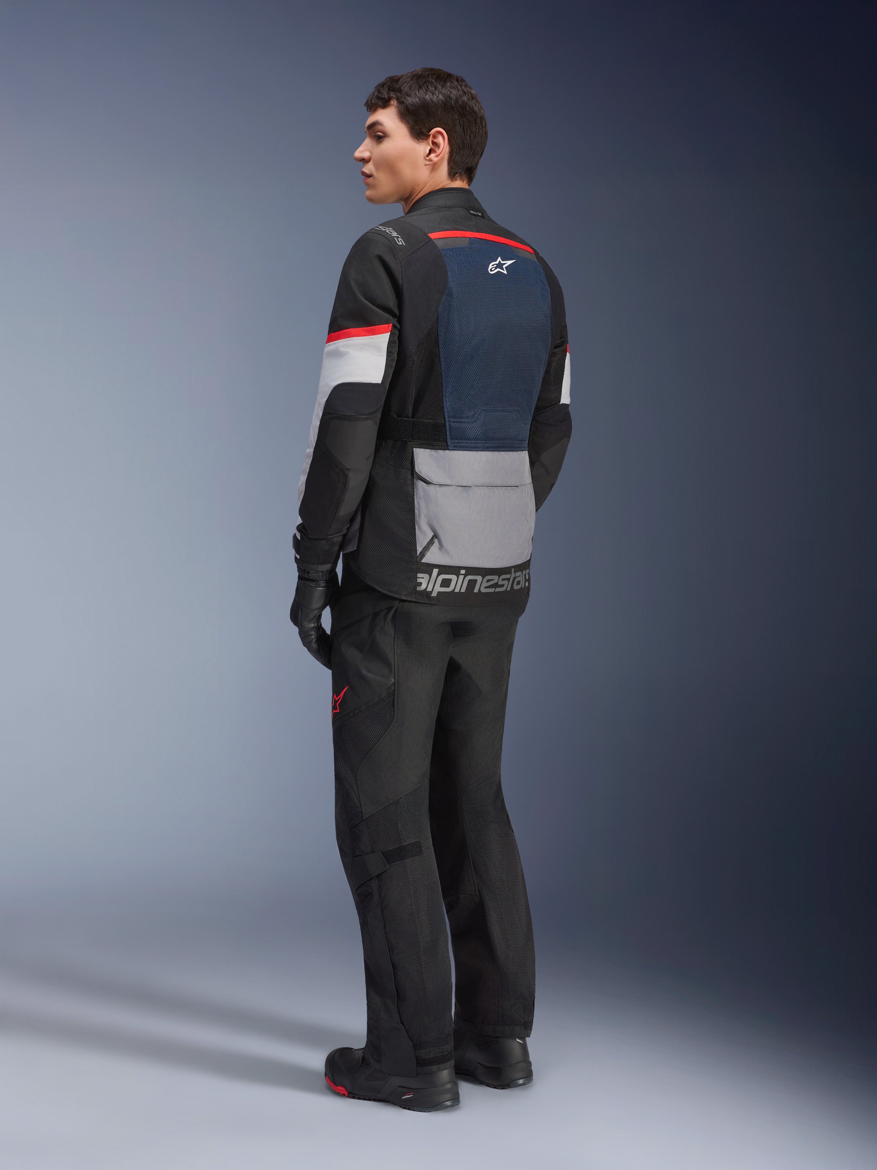 Męski model w ujęciu z tyłu i z boku, ubrany w kurtkę Alpinestars Andes Air Drystar® w kolorze Deep Blue Black Ice Gray, czarne spodnie motocyklowe i rękawice, niebieski panel siatkowy na plecach, szara kieszeń cargo na dole pleców i czerwone akcenty na rękawach, profesjonalna odzież adventure touring.