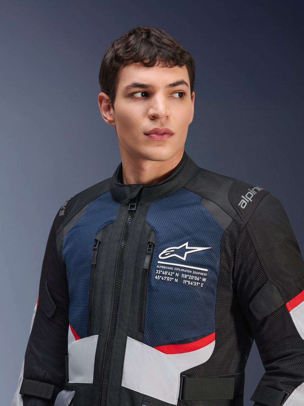 Kurtka Alpinestars Andes Air Drystar®, kurtka motocyklowa, granatowo-czarna i jasnoszara z czerwonymi akcentami, prezentowana na modelu, z wentylowanymi panelami siatkowymi na klatce piersiowej, białym logo gwiazdy ze współrzędnymi geograficznymi i konstrukcją typu adventure.