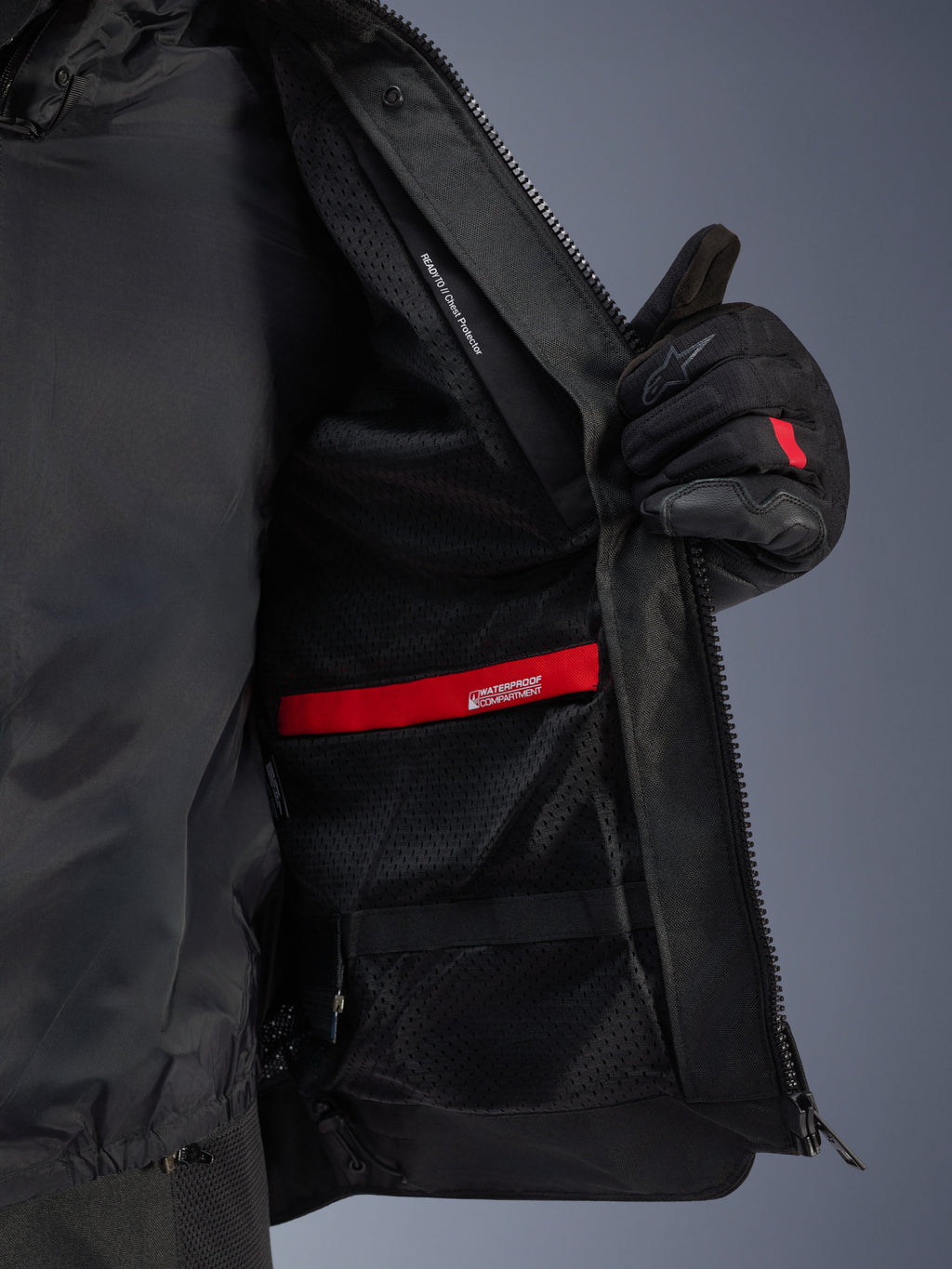 Zbliżenie na wnętrze kurtki Alpinestars Andes Air Drystar®, ukazujące oddychającą czarną podszewkę z siatki, czerwoną wodoodporną przegrodę oraz kieszeń na ochraniacz klatki piersiowej, przytrzymywaną przez czarną rękawicę motocyklową.