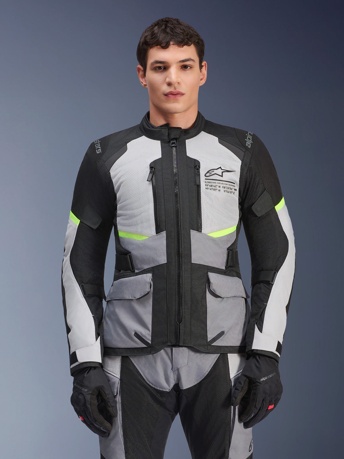 Alpinestars Andes Air Drystar Jacket, turystyczna kurtka motocyklowa adventure, jasnoszara i ciemnoszara z czarnymi panelami i neonowożółtymi akcentami, noszona z dopasowanymi spodniami i rękawicami, z siatkowymi panelami wentylacyjnymi i kieszeniami cargo