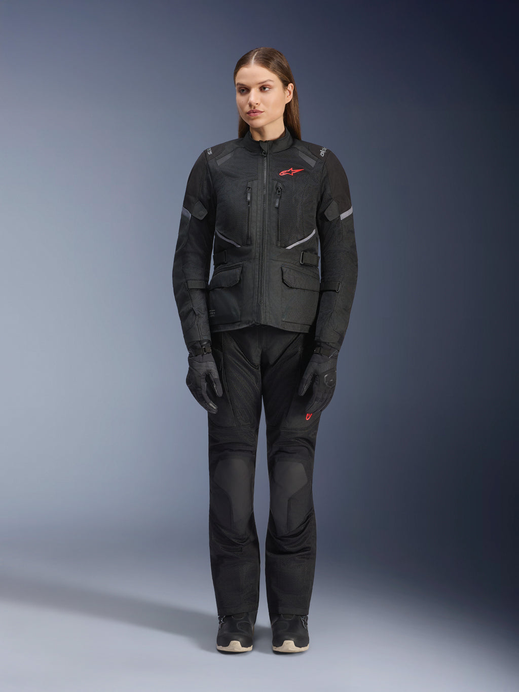 Modelka w czarnej kurtce Alpinestars Stella Andes Air Drystar®, zestawionej z dopasowanymi czarnymi spodniami turystycznymi i rękawicami, z dużymi panelami wentylacyjnymi z siatki, czerwonymi akcentami logo i elementami odblaskowymi, profesjonalne zdjęcie studyjne damskiej odzieży motocyklowej.
