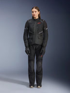 Modelka w czarnej kurtce Alpinestars Stella Andes Air Drystar®, zestawionej z dopasowanymi czarnymi spodniami turystycznymi i rękawicami, z dużymi panelami wentylacyjnymi z siatki, czerwonymi akcentami logo i elementami odblaskowymi, profesjonalne zdjęcie studyjne damskiej odzieży motocyklowej.