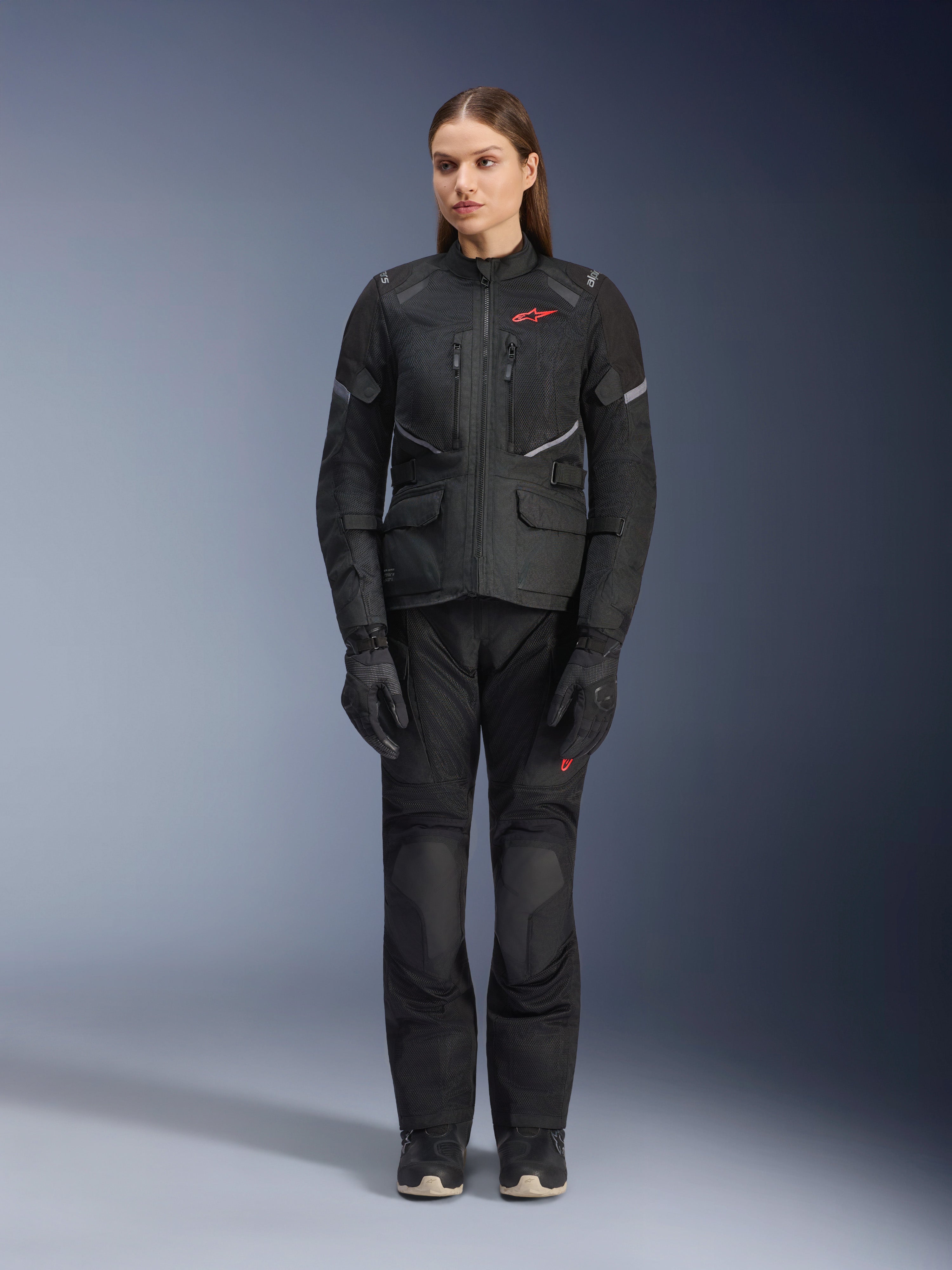 Modelka w czarnej kurtce Alpinestars Stella Andes Air Drystar®, zestawionej z dopasowanymi czarnymi spodniami turystycznymi i rękawicami, z dużymi panelami wentylacyjnymi z siatki, czerwonymi akcentami logo i elementami odblaskowymi, profesjonalne zdjęcie studyjne damskiej odzieży motocyklowej.
