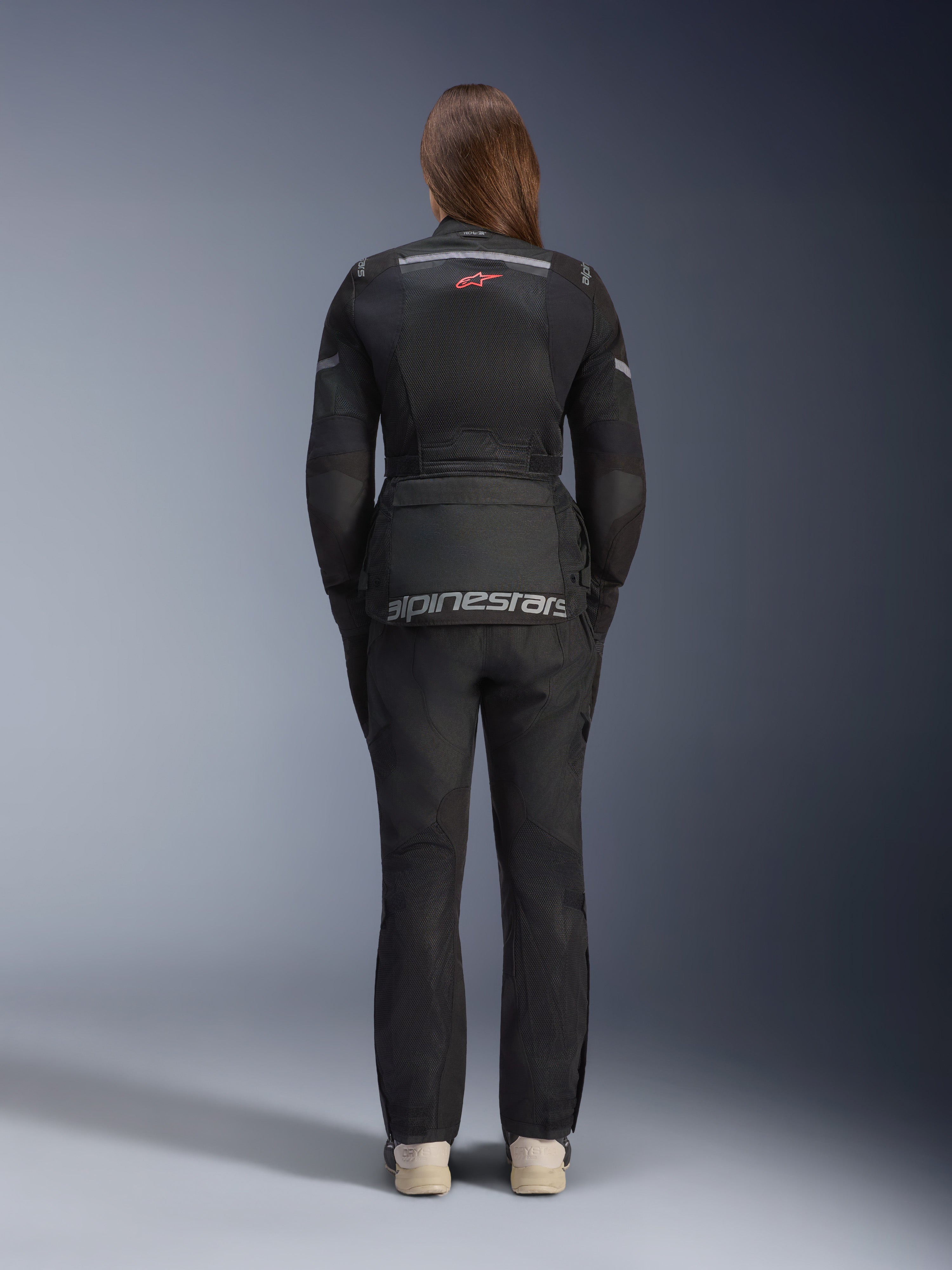 Kobieta ubrana w kurtkę Alpinestars Stella Andes Air Drystar® i pasujące spodnie, kolor czarny, widok z tyłu prezentujący duże szare logo Alpinestars na dolnej kieszeni, małe czerwone logo z gwiazdą na górze pleców, siatkowe panele wentylacyjne i odblaskowe detale do adventure touring