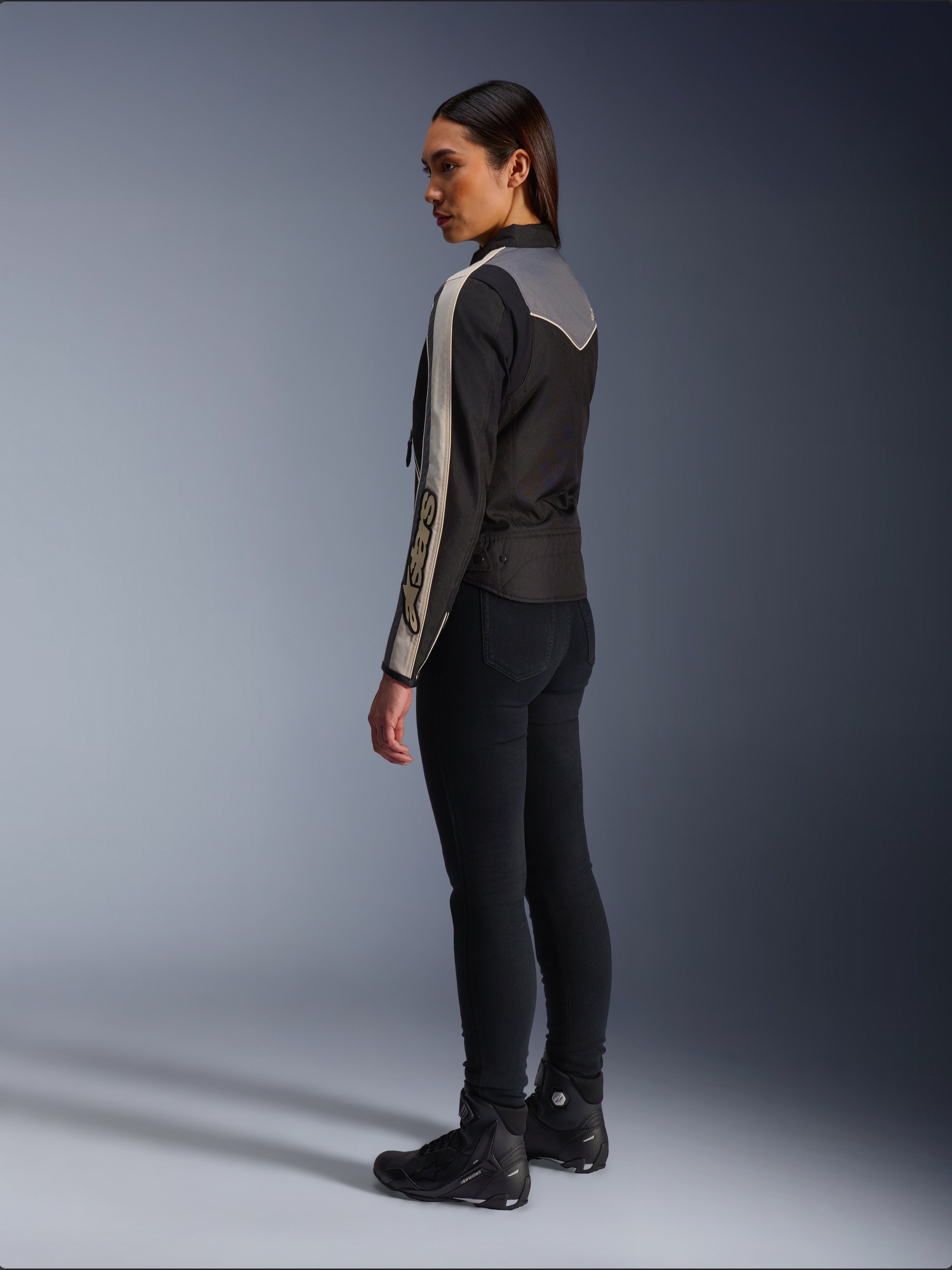 Modelka widziana z boku i z tyłu w kurtce Alpinestars Stella T-Dyno WR w kolorze Black Dark Gray Ecru, z designem inspirowanym stylem vintage, pasami ecru na rękawach i szarym panelem na plecach, zestawiona z czarnymi spodniami i czarnymi butami motocyklowymi.