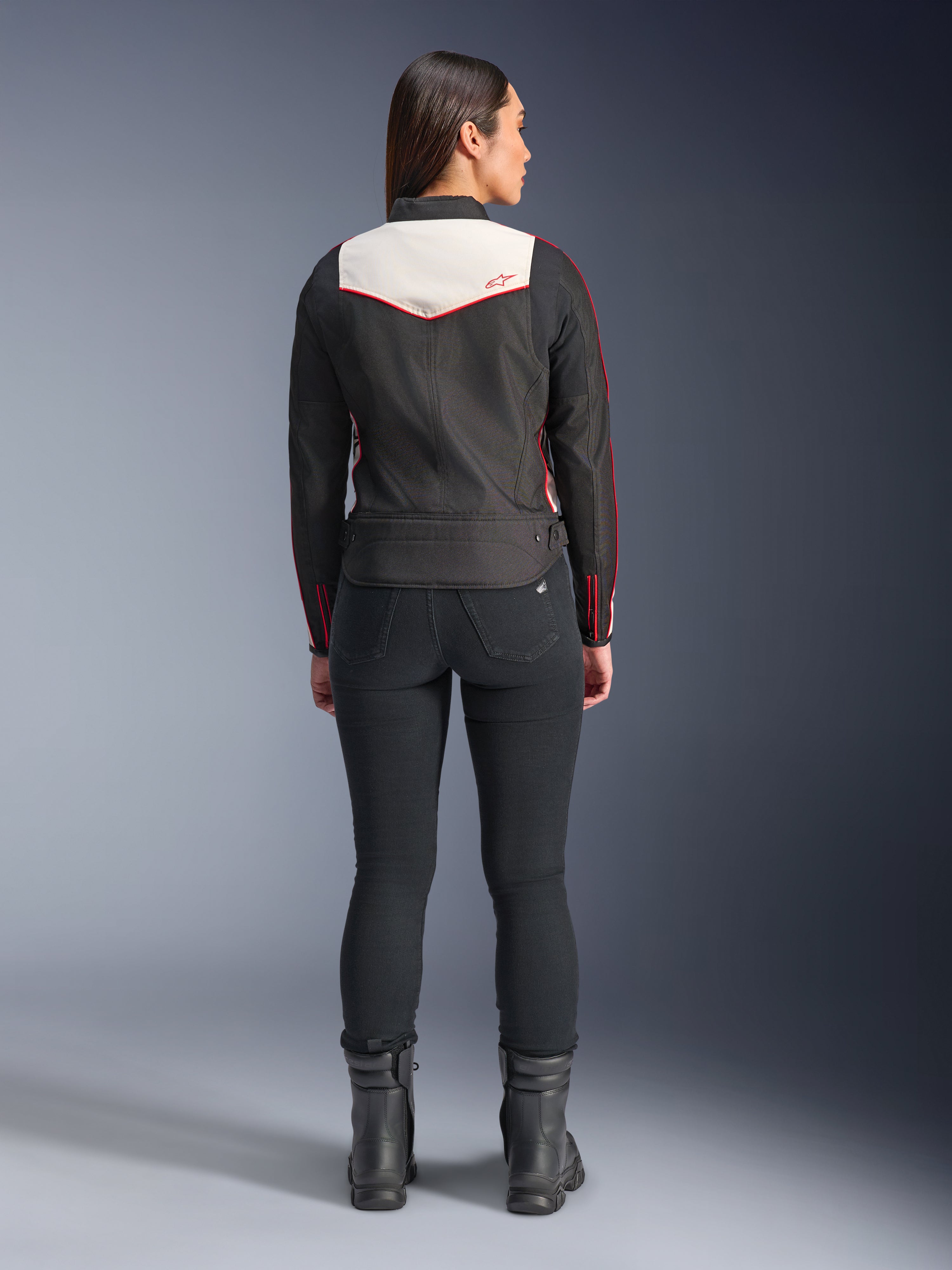 Widok z tyłu modelki ubranej w kurtkę Alpinestars Stella T-Dyno WR w kolorystyce Black Ecru Bright Red, z charakterystycznym kremowym panelem na plecach i czerwonymi akcentami logo, w zestawieniu z czarnymi spodniami i butami, tekstylna kurtka motocyklowa w stylu vintage.
