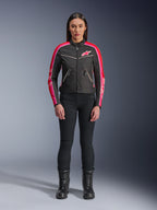 Kobieta ubrana w kurtkę Alpinestars Stella T-Dyno WR, czarną z jaskrawoczerwonymi i fuksjowymi akcentami, tekstylna kurtka w stylu vintage z różowym logo i paskami na rękawach, zestawiona z czarnymi spodniami i sznurowanymi butami, miejska odzież motocyklowa