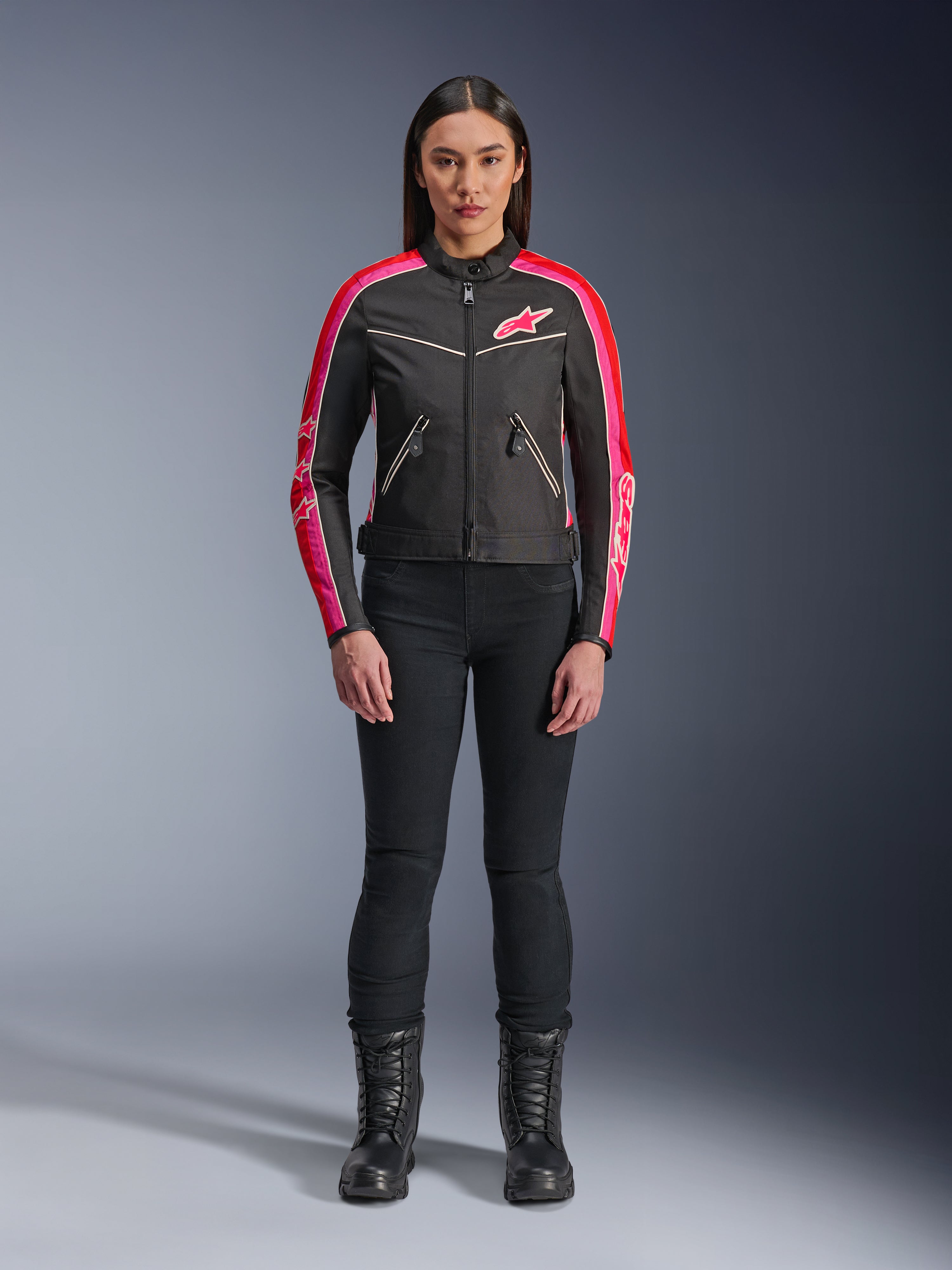 Kobieta ubrana w kurtkę Alpinestars Stella T-Dyno WR, czarną z jaskrawoczerwonymi i fuksjowymi akcentami, tekstylna kurtka w stylu vintage z różowym logo i paskami na rękawach, zestawiona z czarnymi spodniami i sznurowanymi butami, miejska odzież motocyklowa