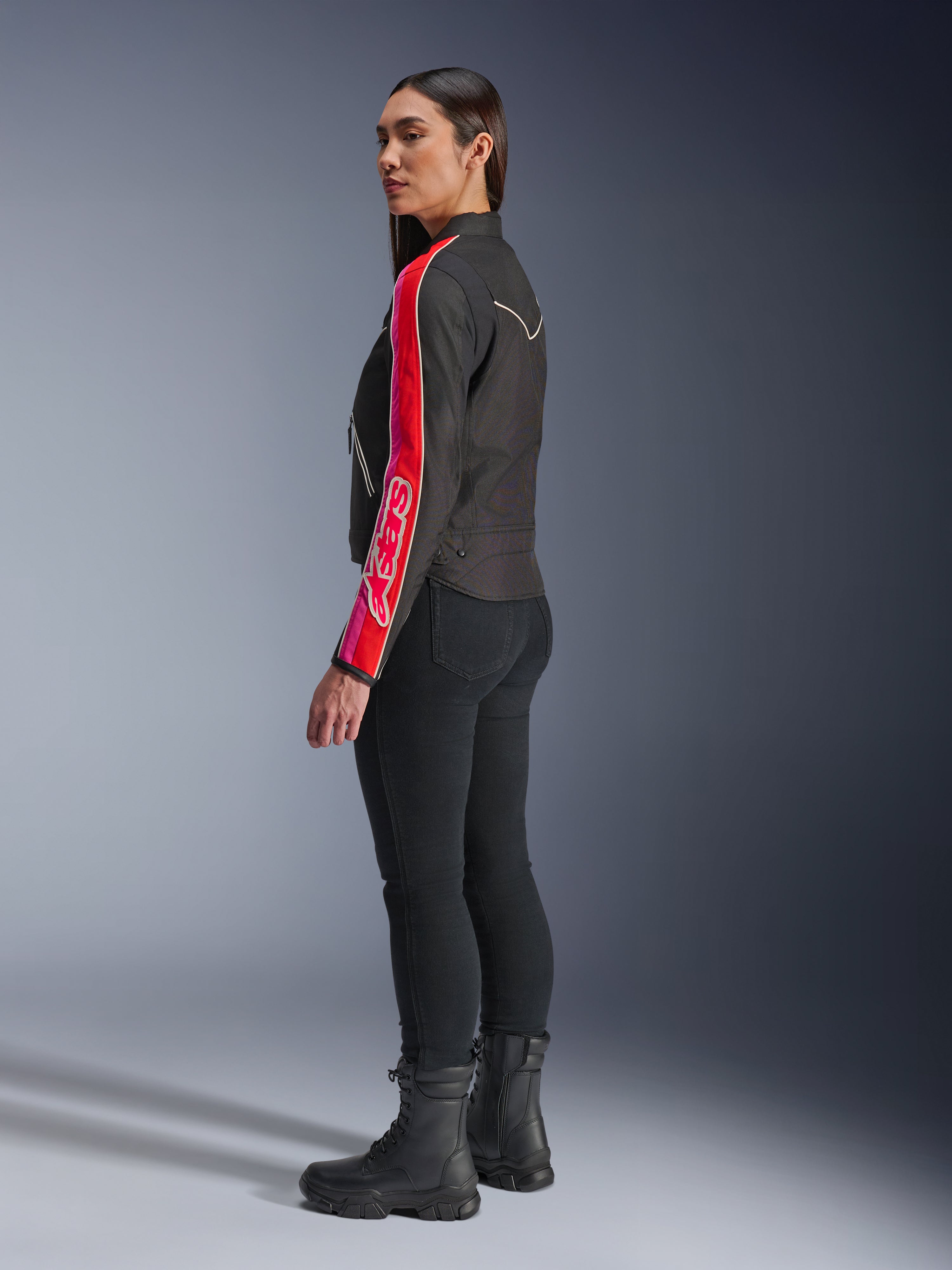 Modelka ubrana w kurtkę Alpinestars Stella T-Dyno WR w kolorze Black Bright Red Fuchsia, z wyrazistymi czerwonymi i różowymi pasami oraz logo Alpinestars na rękawie, zestawiona z czarnymi spodniami do jazdy i czarnymi butami motocyklowymi w studiu.
