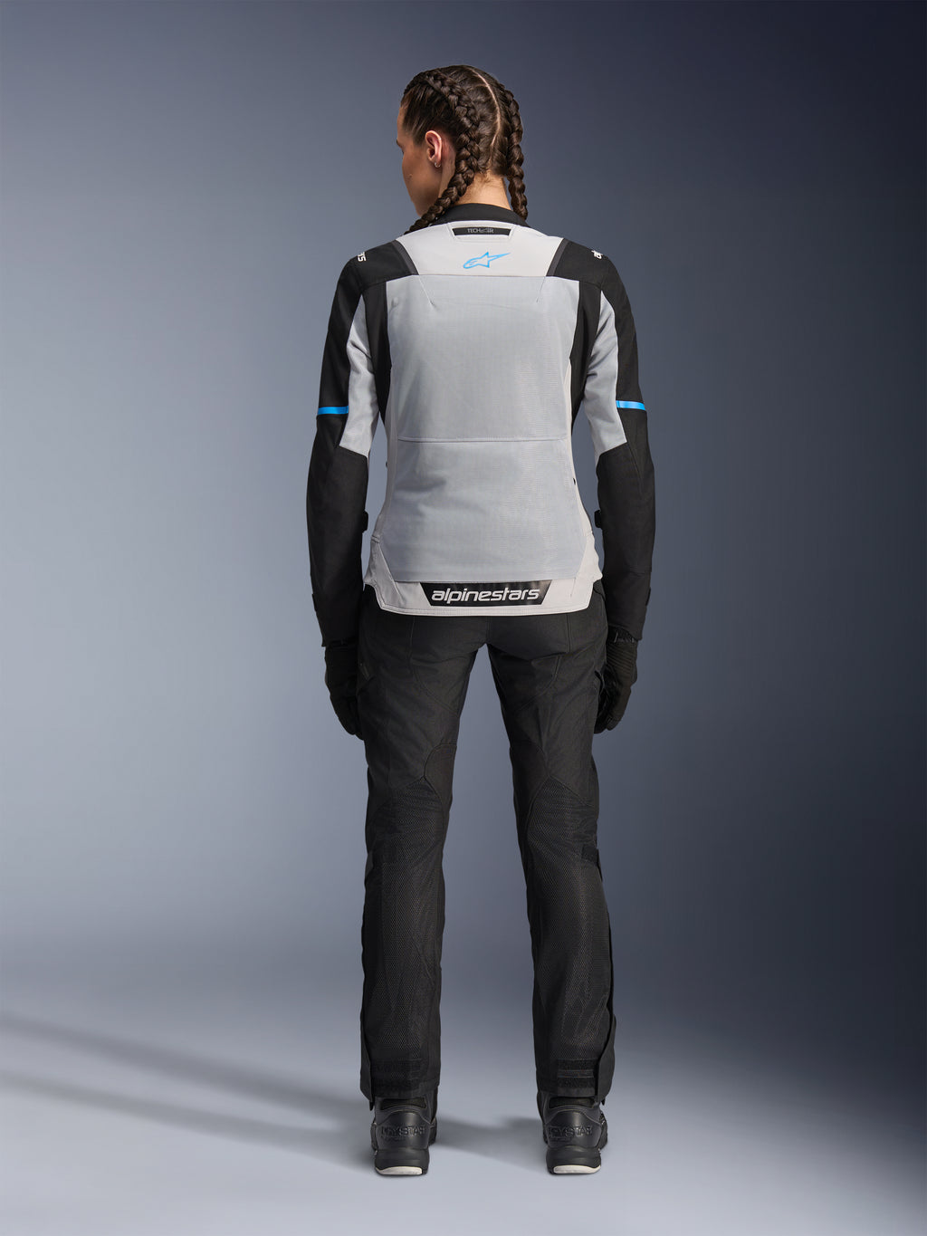 Damska kurtka Alpinestars Stella ST-2 Air, kolor Ice Gray Black, widok z tyłu na modelce, duży jasnoszary siatkowy panel na plecach, czarne rękawy z niebieskimi akcentami, logo Alpinestars na dole pleców, czarne techniczne spodnie i buty motocyklowe, letnia odzież turystyczna