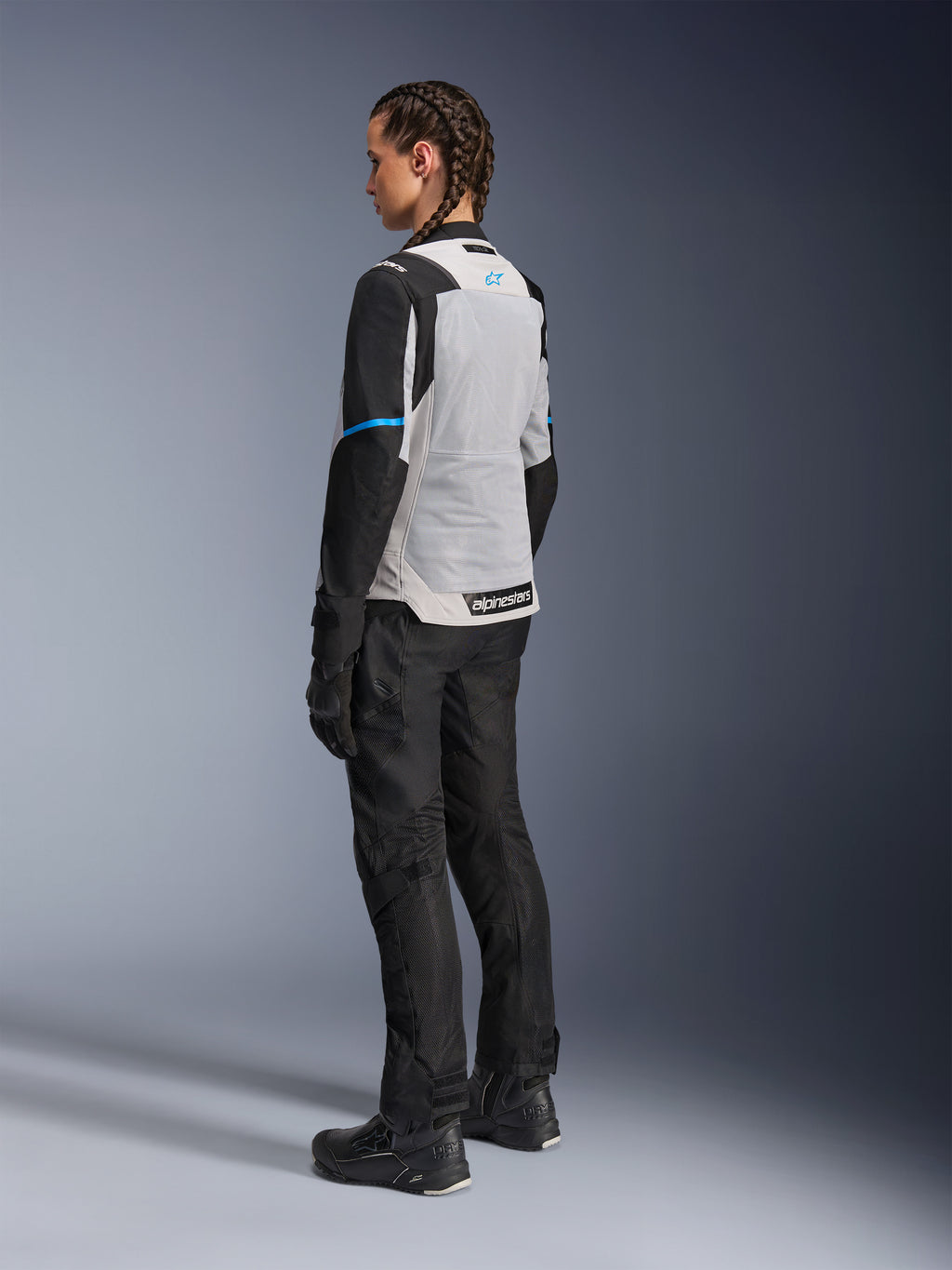 Motocyklistka ubrana w kurtkę Alpinestars Stella ST-2 Air w kolorze Ice Gray i czarnym, widok z tyłu pod kątem trzech czwartych podkreślający duży jasnoszary panel siatkowy na plecach, czarne rękawy z niebieskim paskiem i niebieskim logo Alpinestars, zestawiona z czarnymi spodniami motocyklowymi, rękawicami i butami, tworząca kompletny strój do turystyki przygodowej