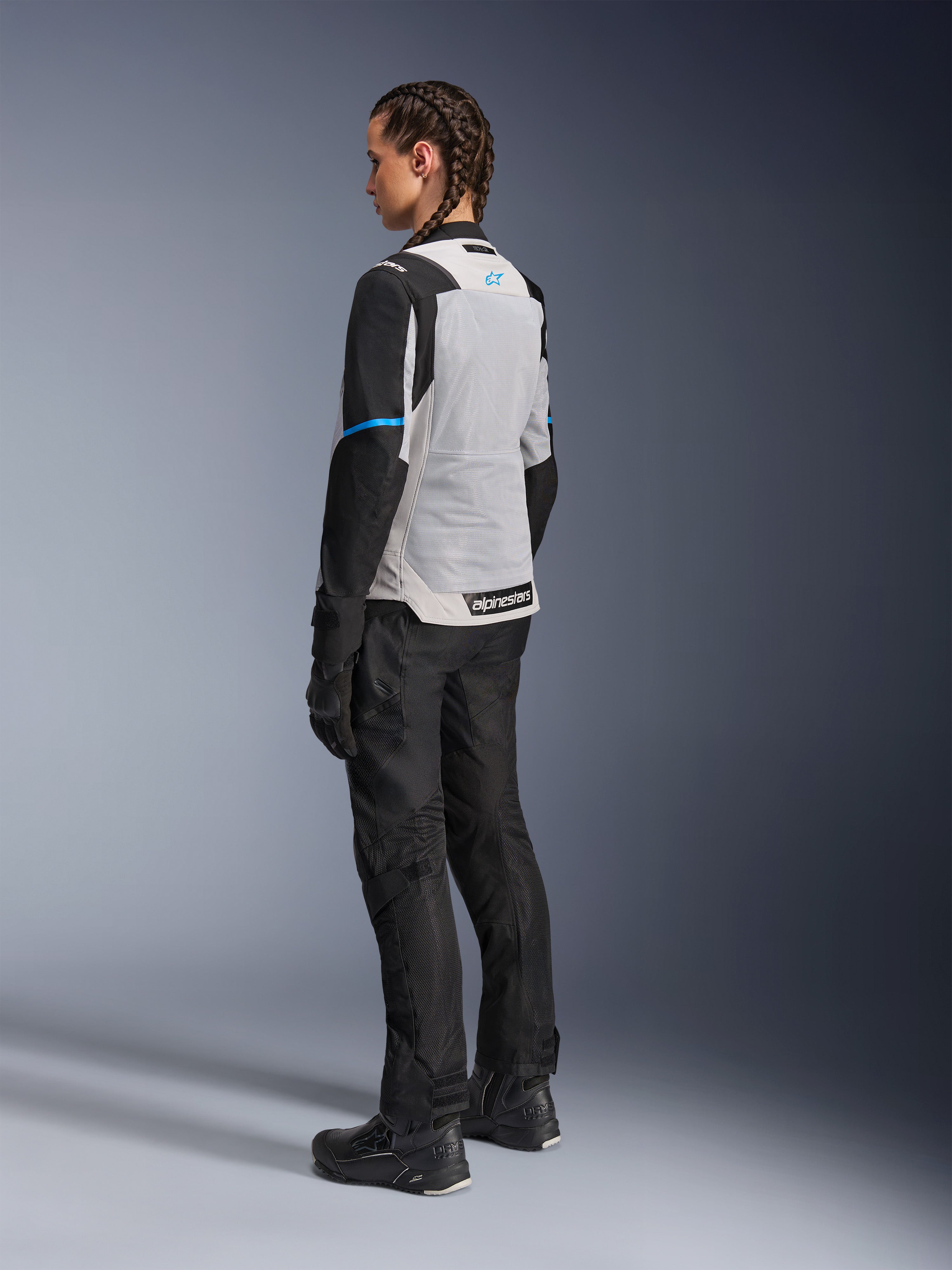 Motocyklistka ubrana w kurtkę Alpinestars Stella ST-2 Air w kolorze Ice Gray i czarnym, widok z tyłu pod kątem trzech czwartych podkreślający duży jasnoszary panel siatkowy na plecach, czarne rękawy z niebieskim paskiem i niebieskim logo Alpinestars, zestawiona z czarnymi spodniami motocyklowymi, rękawicami i butami, tworząca kompletny strój do turystyki przygodowej
