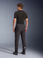 Spodnie Alpinestars Aeroshell, czarne, motocyklowe spodnie softshellowe, widok z tyłu na modelu, o nowoczesnym, zwężanym kroju, białe logo Astars na dole nogawki, elastyczne ściągacze, noszone z czarną warstwą bazową z krótkim rękawem i butami motocyklowymi.