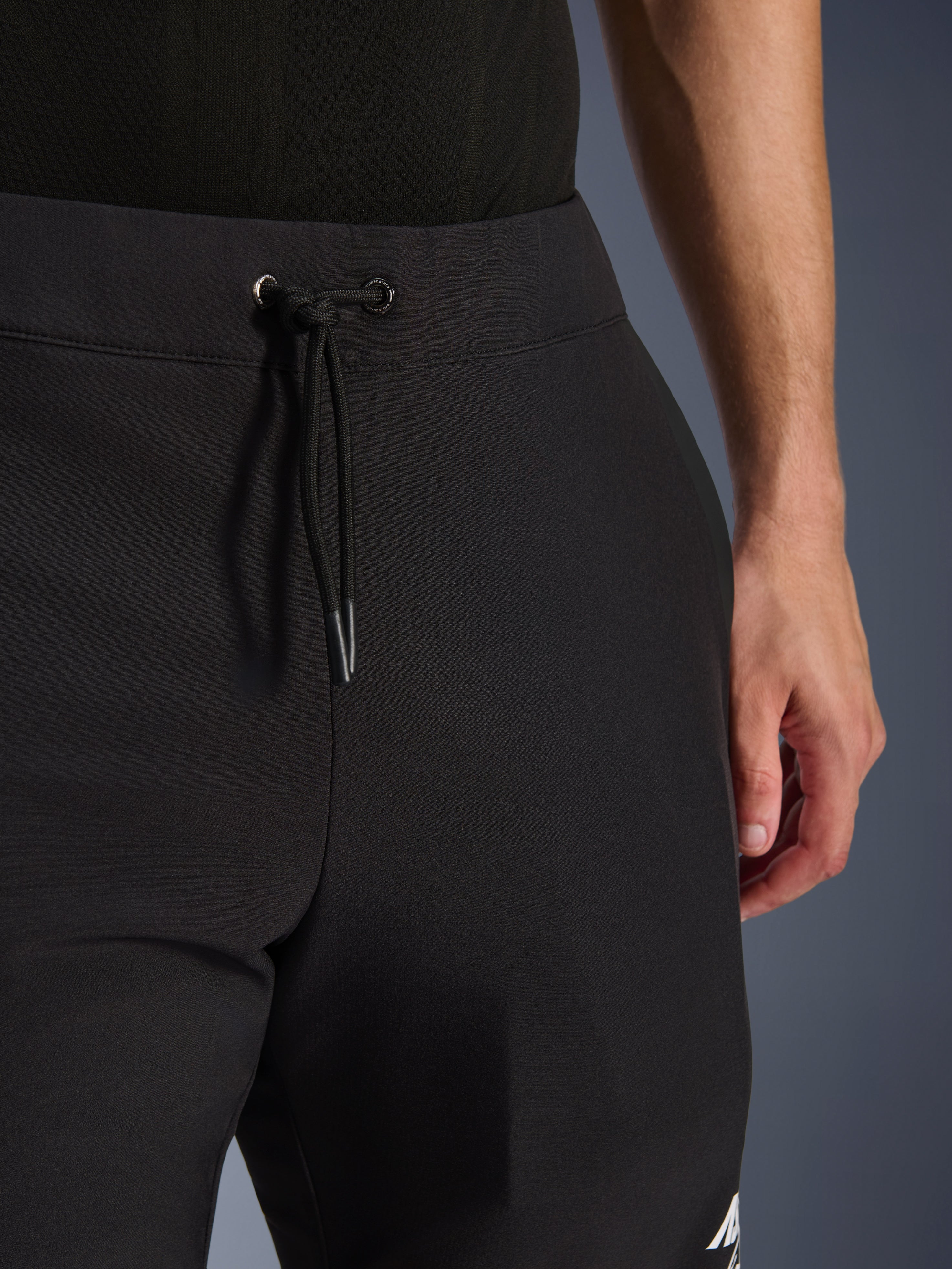 Alpinestars Aeroshell Pants, czarne motocyklowe spodnie softshellowe, zbliżenie na elastyczny pas i zapięcie na sznurek, nowoczesny minimalistyczny design, wytrzymała konstrukcja tekstylna do casualowej jazdy turystycznej