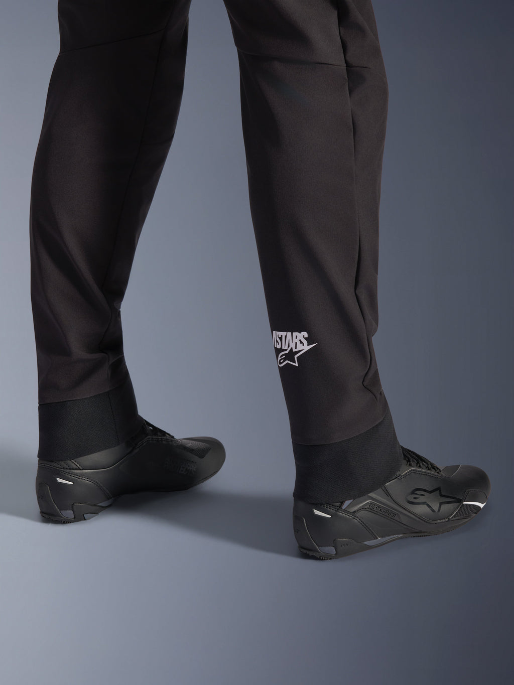 Spodnie Alpinestars Aeroshell, czarne motocyklowe spodnie softshellowe, zbliżenie na dolną część nogawki i elastyczny ściągacz, białe logo Alpinestars na łydce, zestawione z czarnymi butami motocyklowymi, nowoczesny sportowy design typu crossover