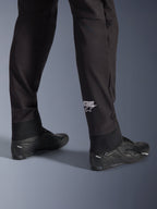 Spodnie Alpinestars Aeroshell, czarne motocyklowe spodnie softshellowe, zbliżenie na dolną część nogawki i elastyczny ściągacz, białe logo Alpinestars na łydce, zestawione z czarnymi butami motocyklowymi, nowoczesny sportowy design typu crossover