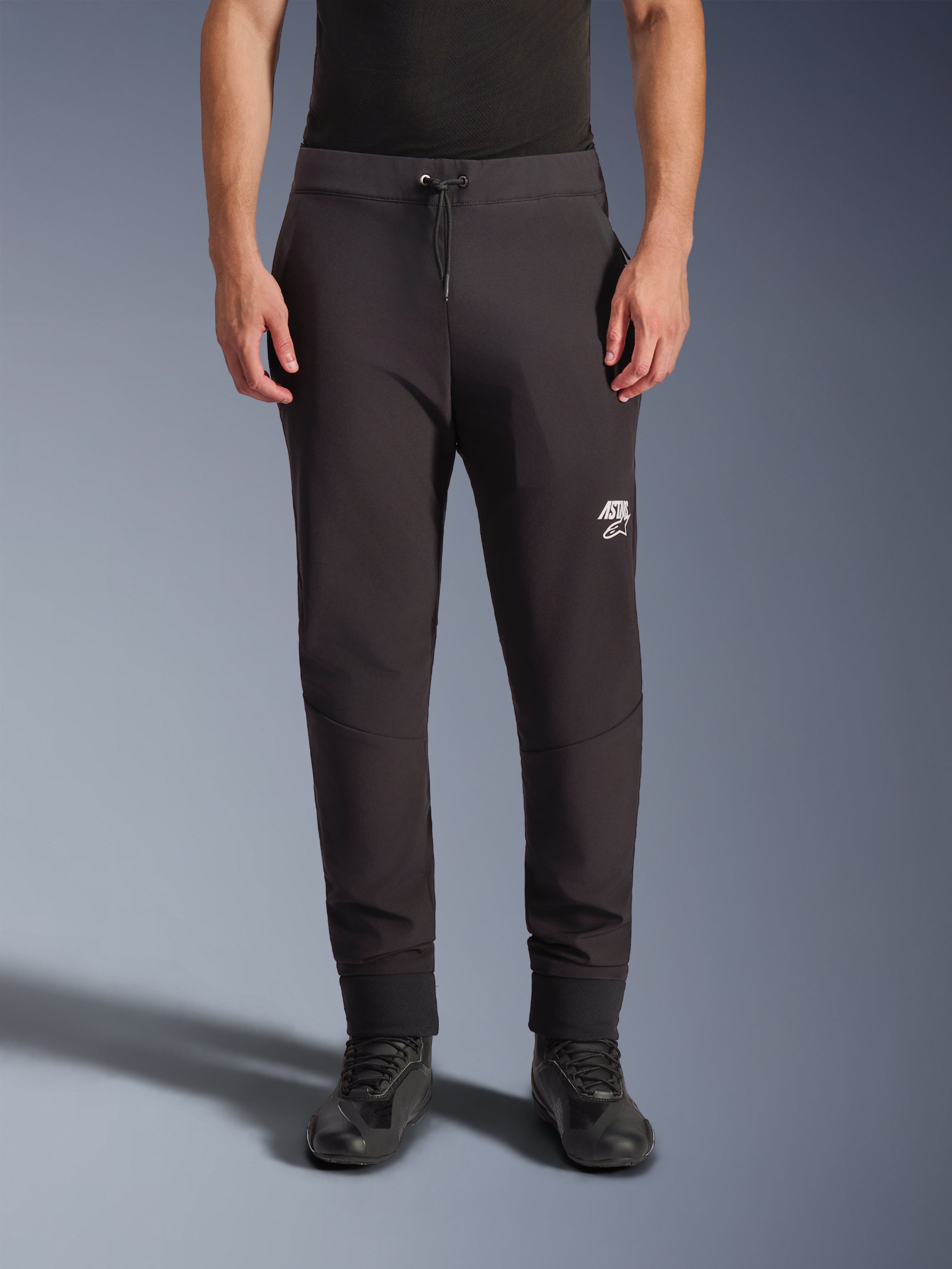 Spodnie Alpinestars Aeroshell, czarne motocyklowe spodnie softshellowe, sportowy krój typu jogger z troczkiem w pasie i elastycznymi mankietami, białe logo Astars, noszone z czarnymi butami motocyklowymi, nowoczesna odzież typu crossover do użytku szosowego i casualowego